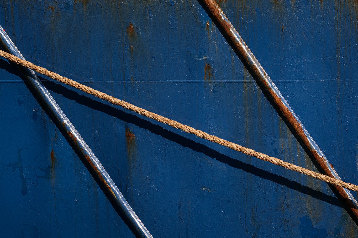 Diagonal ropes and metal pipes creating an abstract pattern across a weathered blue ship hull.