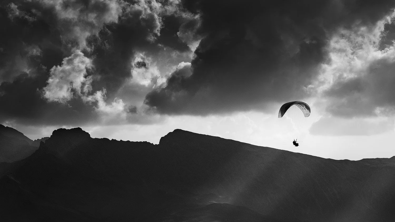 Black-and-white photograph of a paraglider above a silhouetted mountain ridge beneath dramatic clouds at Seceda.