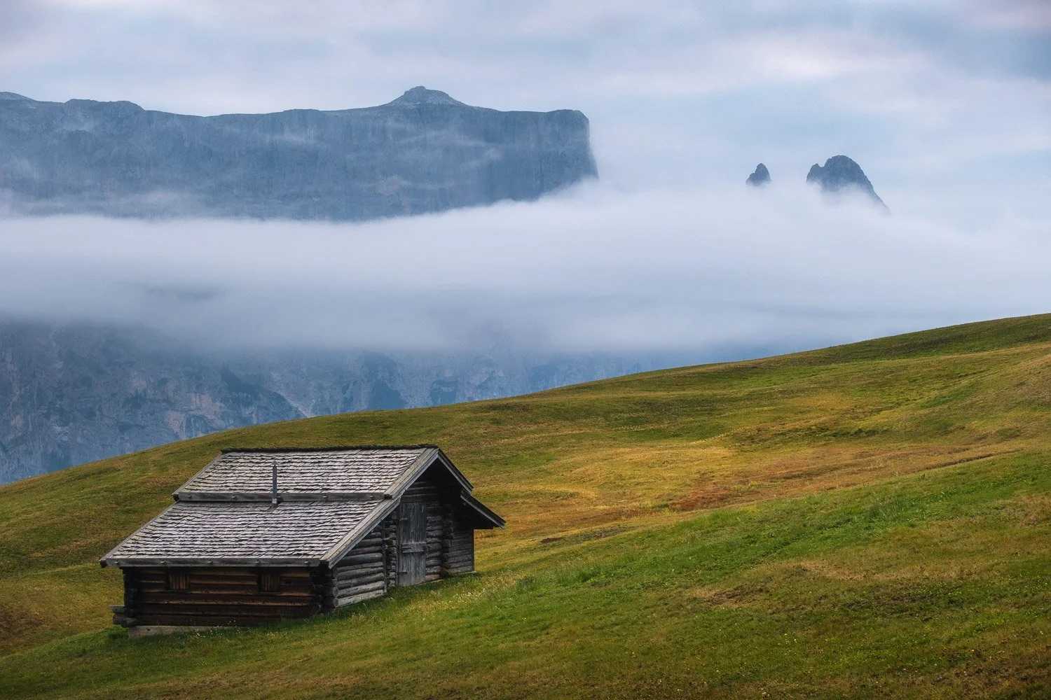 A wooden hut on a green meadow at Alpe di Siusi, with low cloud drifting beneath distant mountain cliffs.