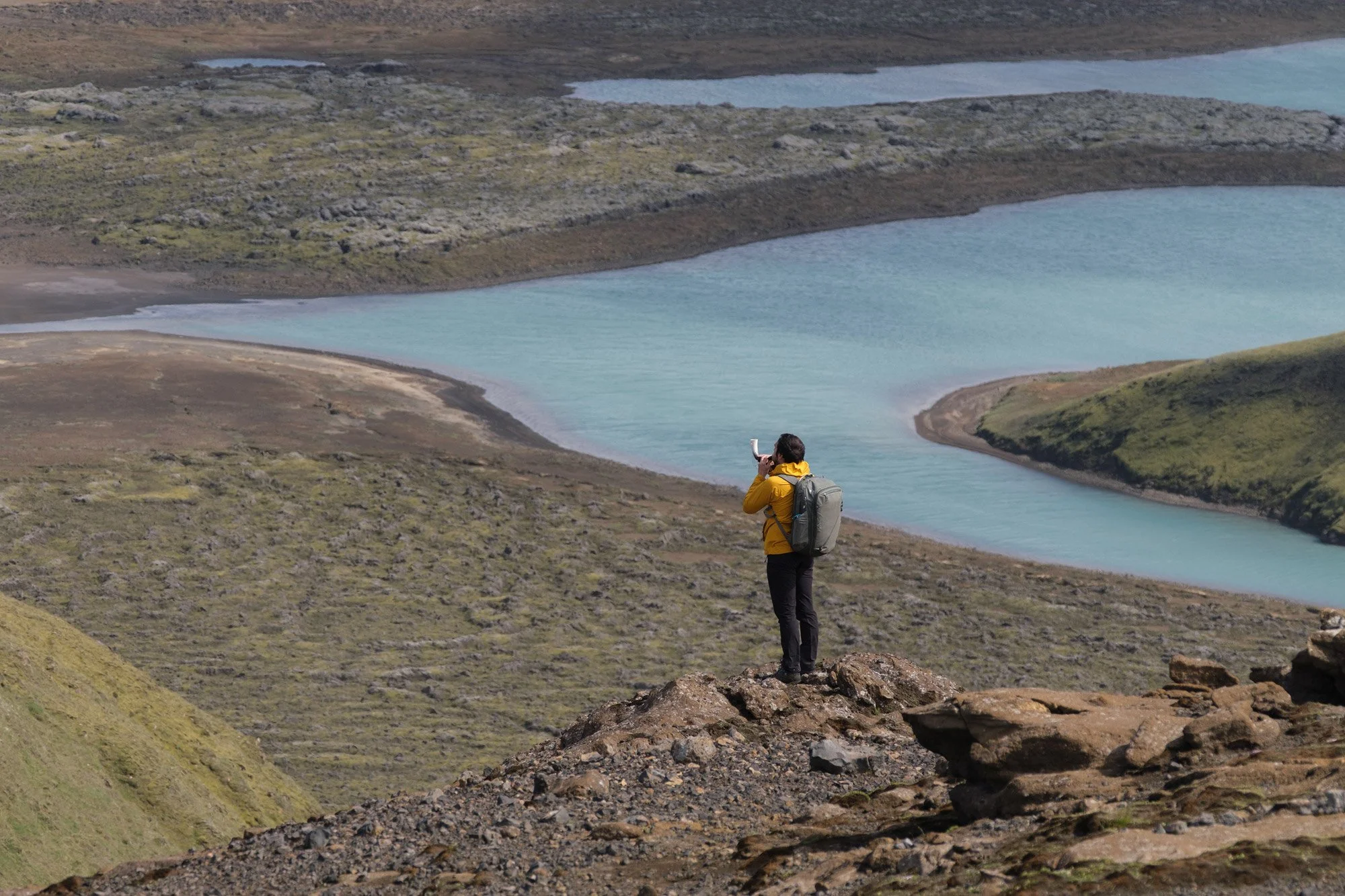 Photographer in a yellow jacket standing on a rocky ridge above turquoise lakes in the Icelandic Highlands, photographed with the Fujinon 50–140mm and 1.4× teleconverter.