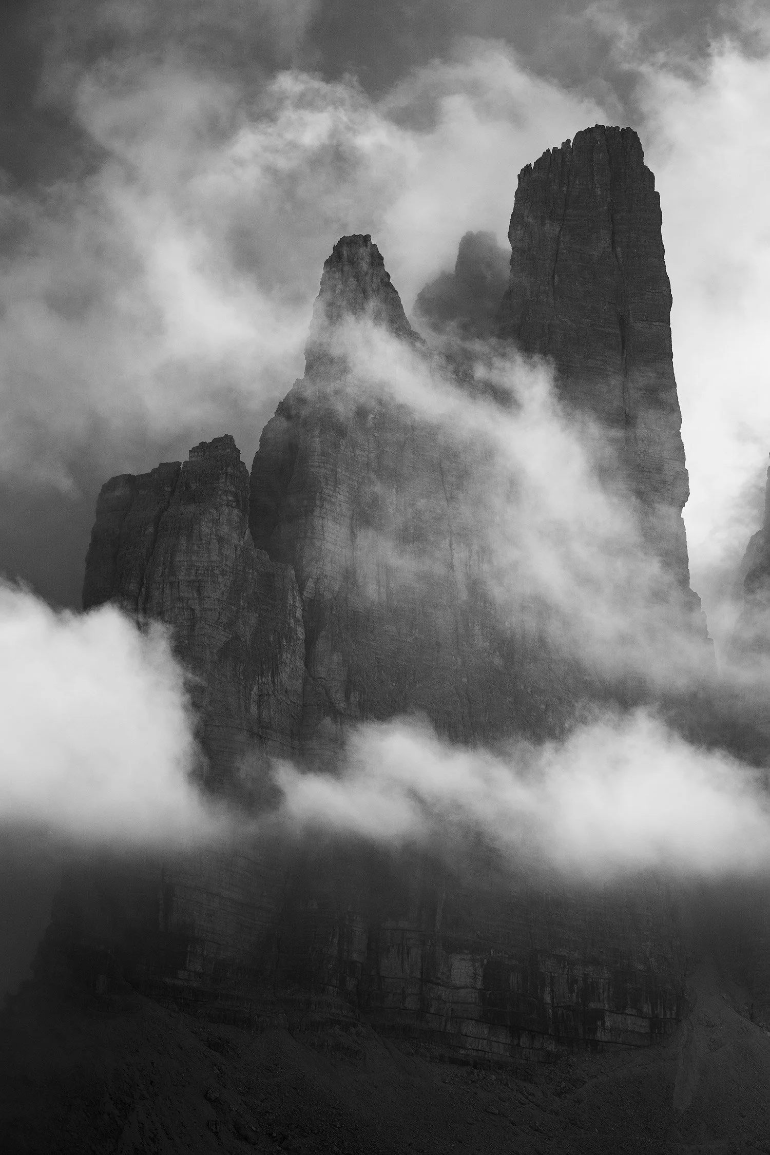 Black-and-white photograph of jagged rock towers emerging from clouds in the Tre Cime di Lavaredo area.