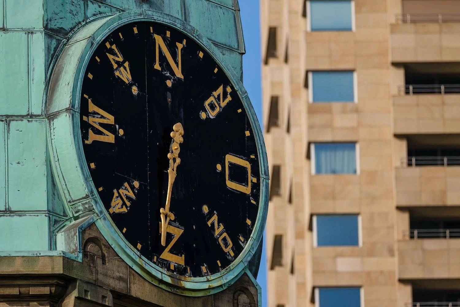 Black-and-gold Roman numeral clock on an oxidized copper tower with apartment buildings behind in Rotterdam.