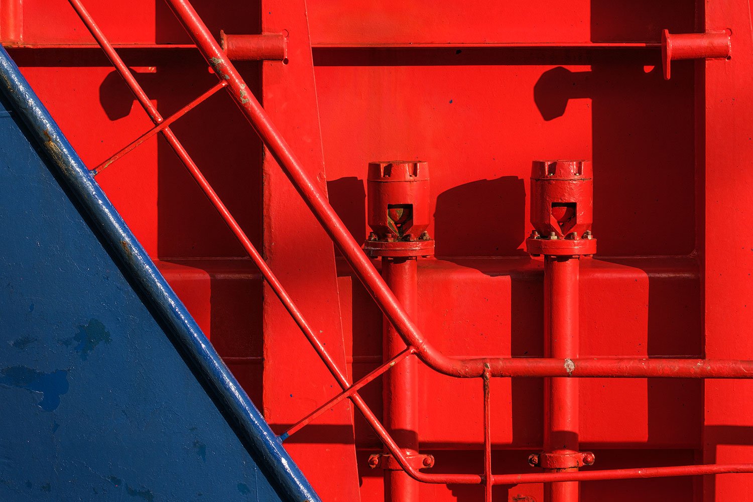 Tight detail of red ship machinery crossed by blue metal rails and pipes.