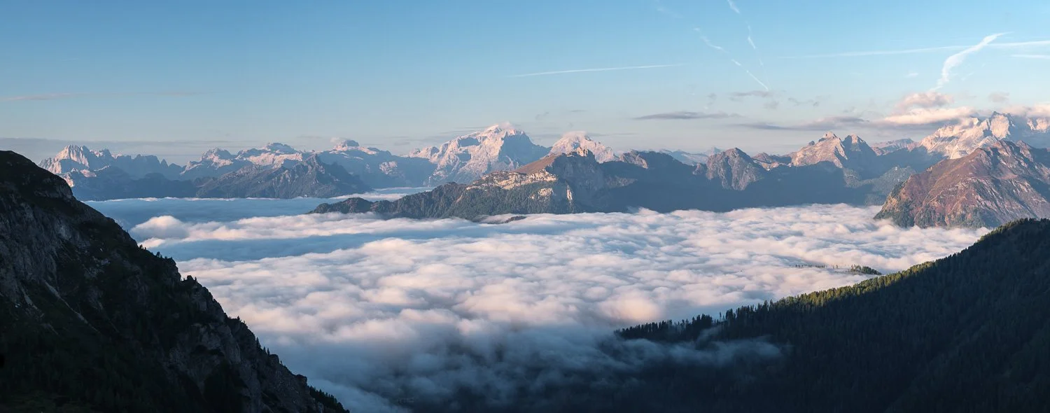 Wide panoramic mountain landscape above a sea of clouds with forested slopes and distant sunlit peaks.