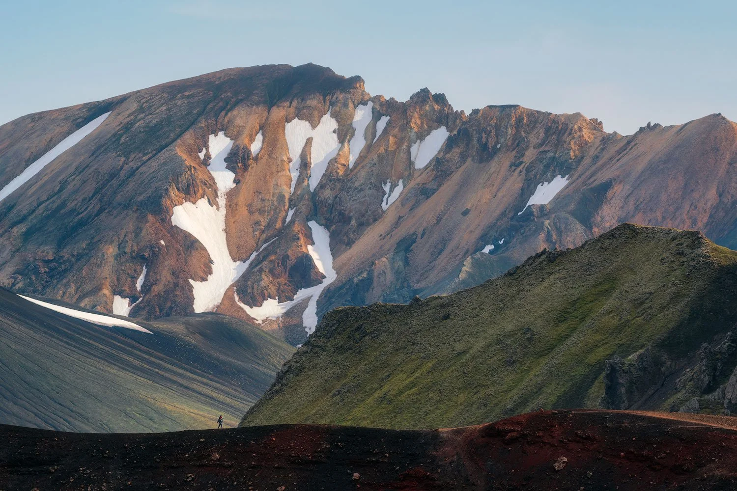 Lone hiker crossing a black volcanic ridge beneath snow-streaked mountains at Landmannalaugar in the Icelandic Highlands.