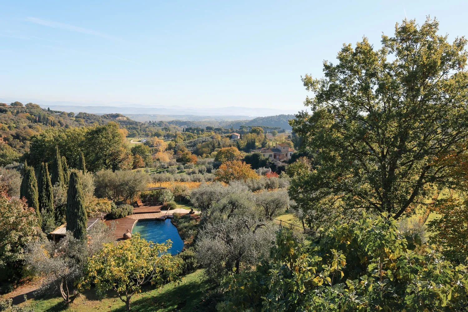 A scenic view of a lush garden with a swimming pool, surrounded by trees, rolling hills, and a few houses under a clear blue sky.