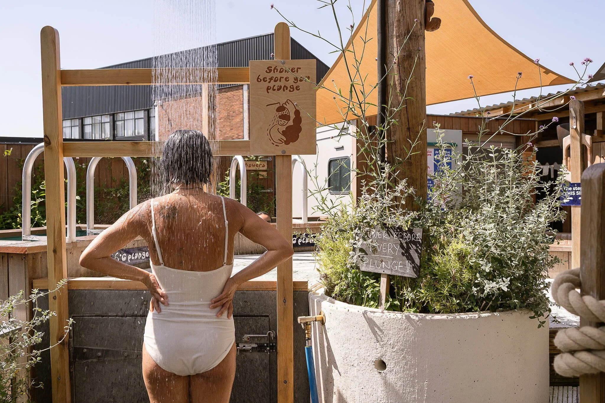 A person is taking an outdoor shower under a wooden structure with a sign that reads "shower before you plunge," with water streaming over their head.