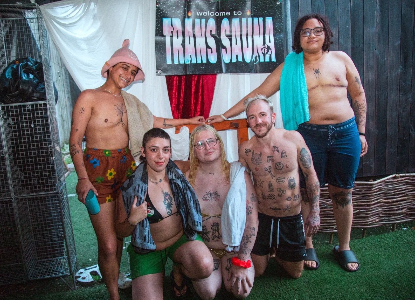 A smiling group in swimwear and towels posing in front of a 'Welcome to Trans Sauna' banner