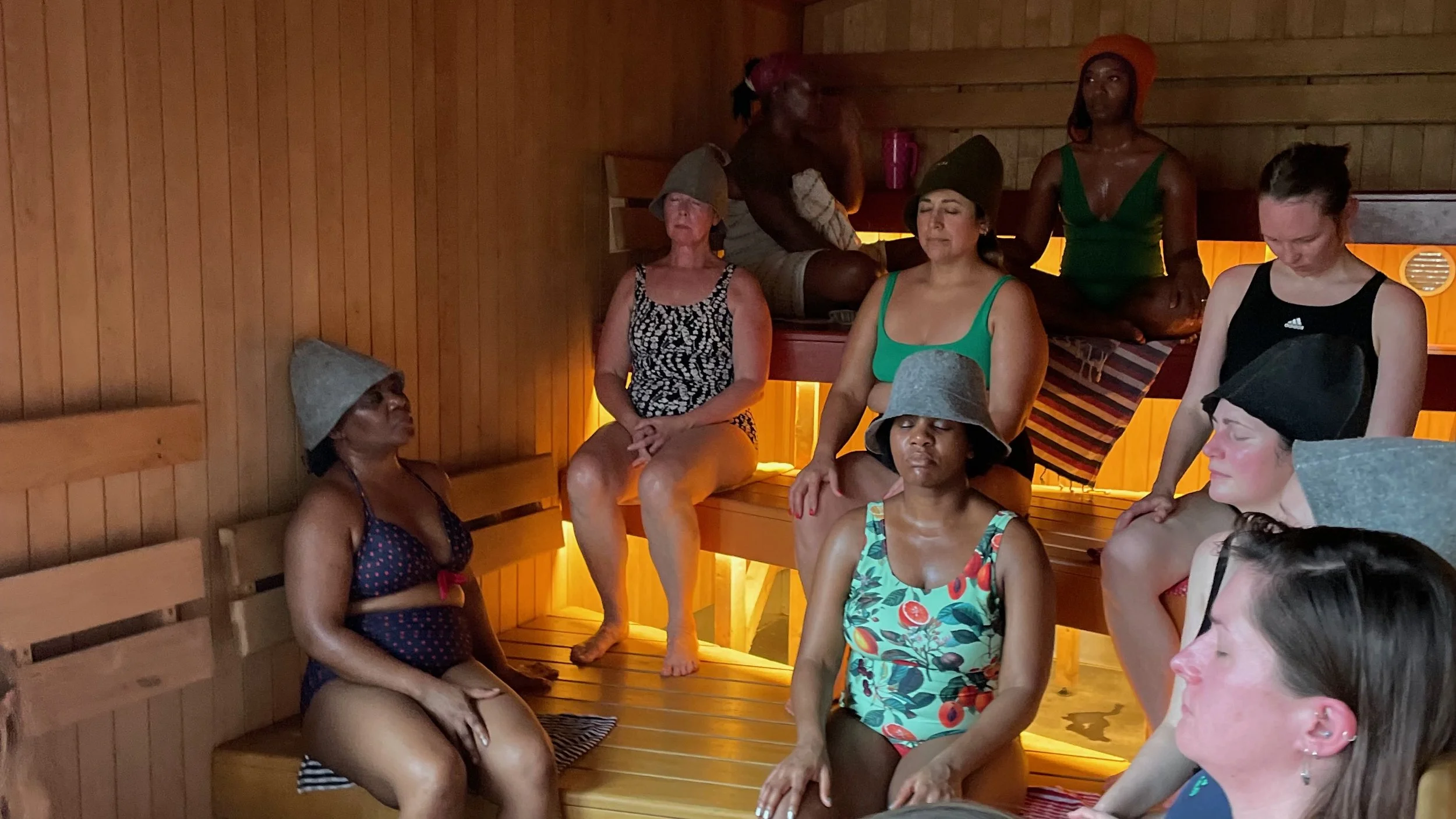 Women in swimsuits and felt sauna hats sitting with their eyes closed on the benches of a wooden sauna
