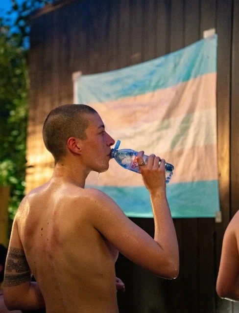 A person with a shaved head drinking from a water bottle in front of a trans pride flag at dusk