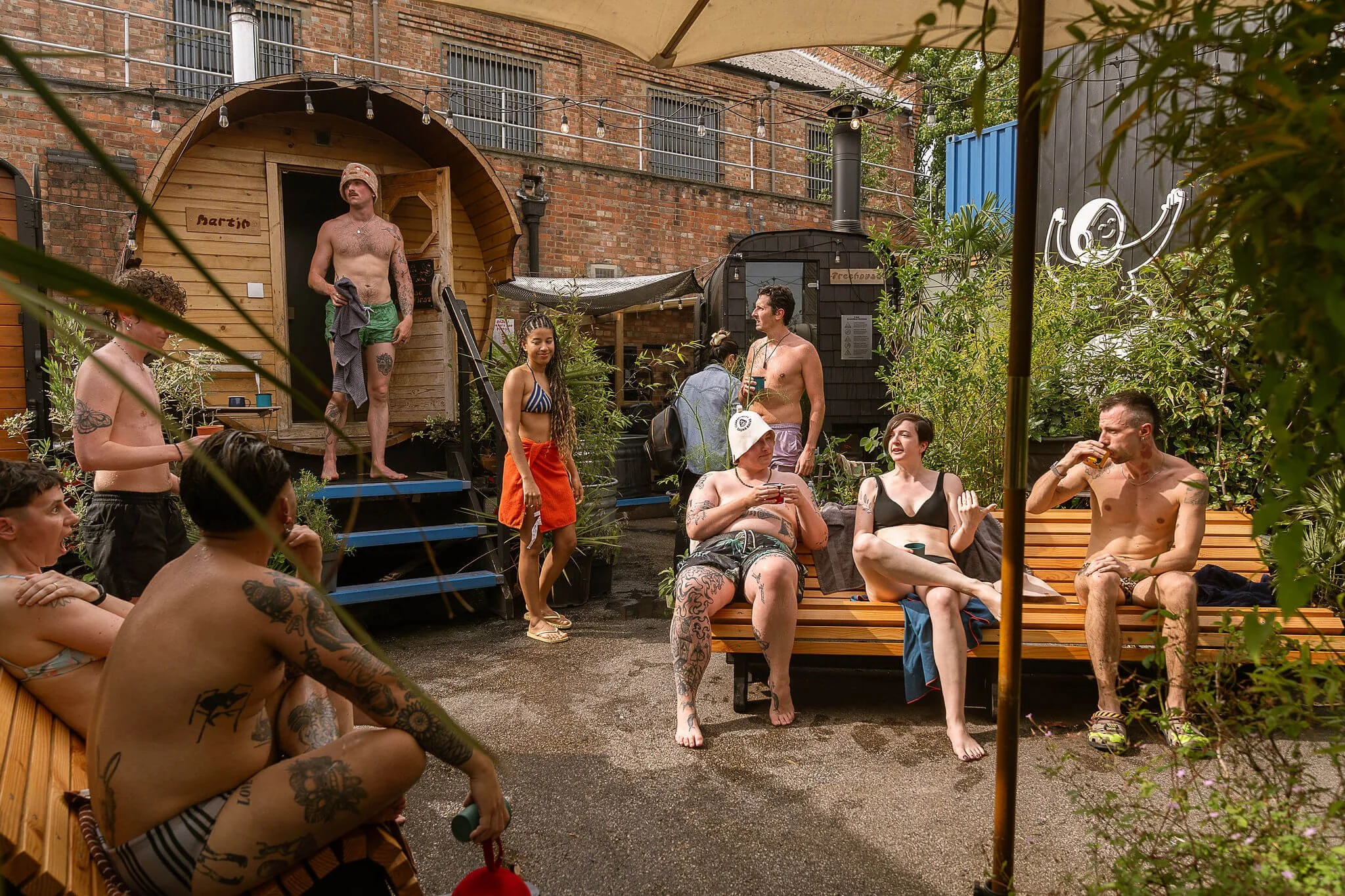 People gathered outdoors in a backyard with a brick building, a small wooden sauna, plants, and string lights. Some individuals are sitting, chatting, and drinking, while others stand or walk around. Many are shirtless or in swimwear, suggesting a casual social gathering or summer party.