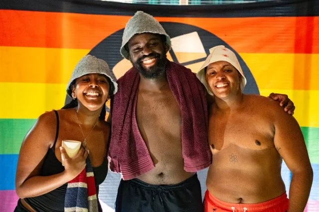 Three people smiling in front of a rainbow flag, two women wearing towels and hats, and a man with a towel around his neck, shirtless.