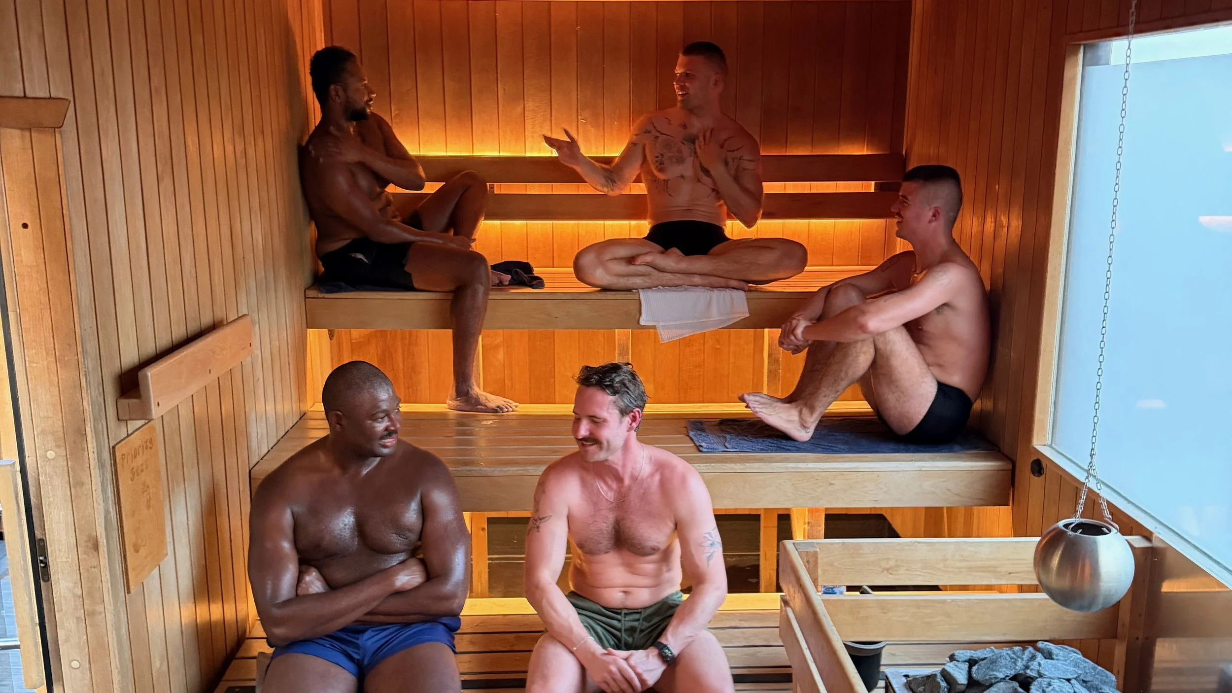 Five men in swim shorts chatting and relaxing on the tiered benches of a warmly lit wooden sauna