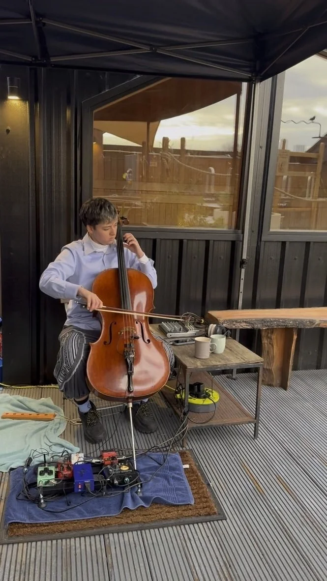 A cellist playing on the outdoor deck by the saunas, with effects pedals at their feet