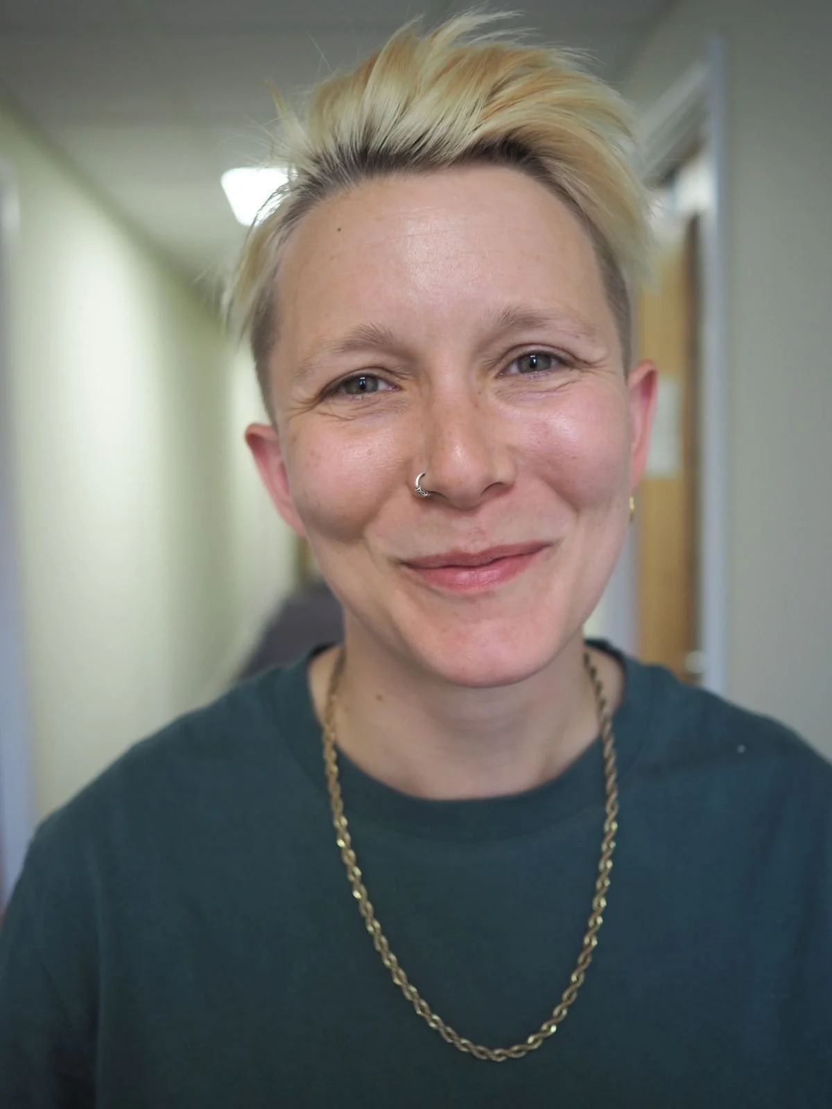 A woman with short blonde hair smiling, wearing a dark shirt, a gold chain necklace, and a nose piercing, in an indoor hallway.