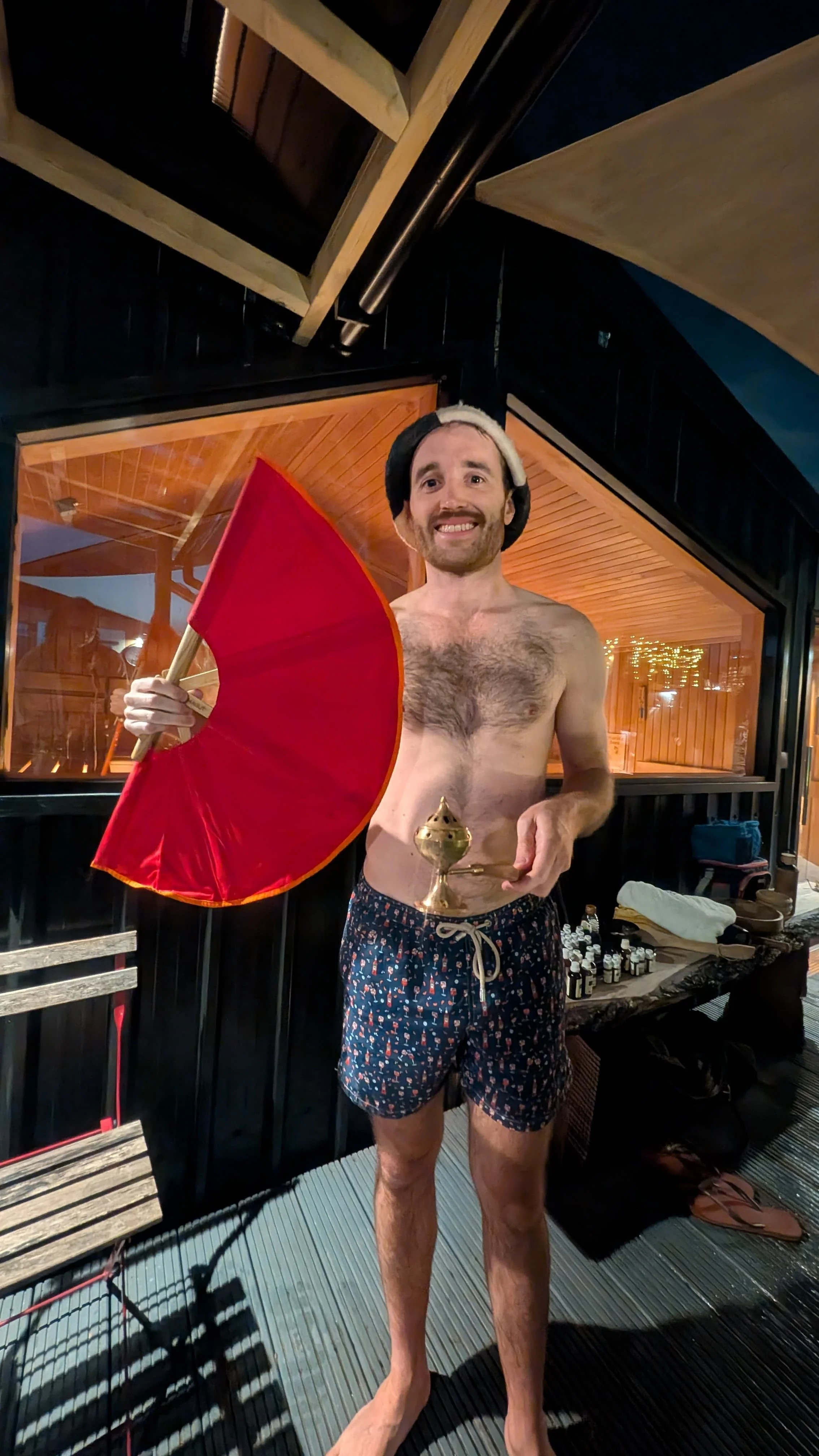 Oliver Wessely smiling in swim shorts and a felt sauna hat, holding a large red fan and a brass incense burner outside a sauna at night