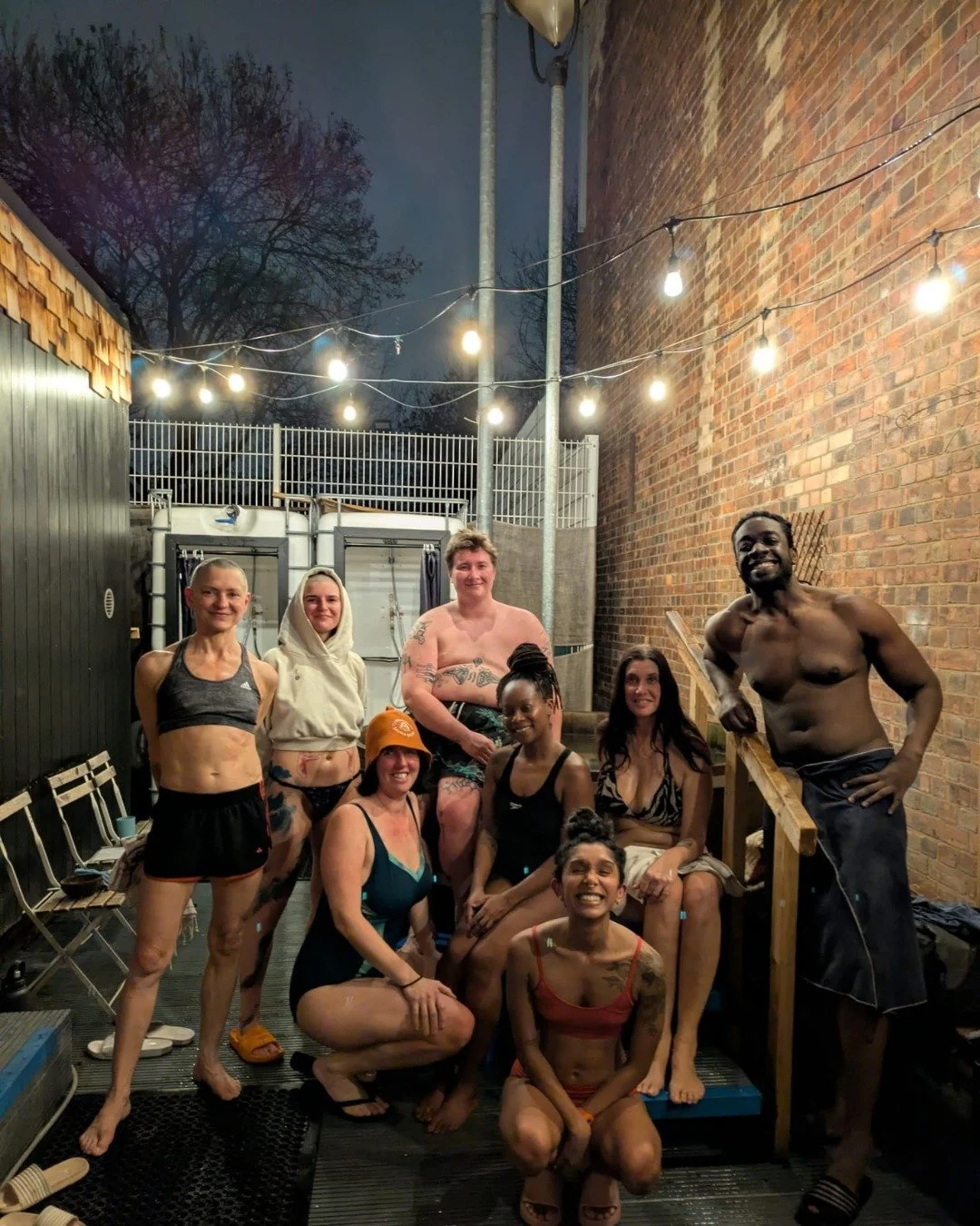 A smiling group in swimwear and towels posing together in an outdoor courtyard at night under festoon lights