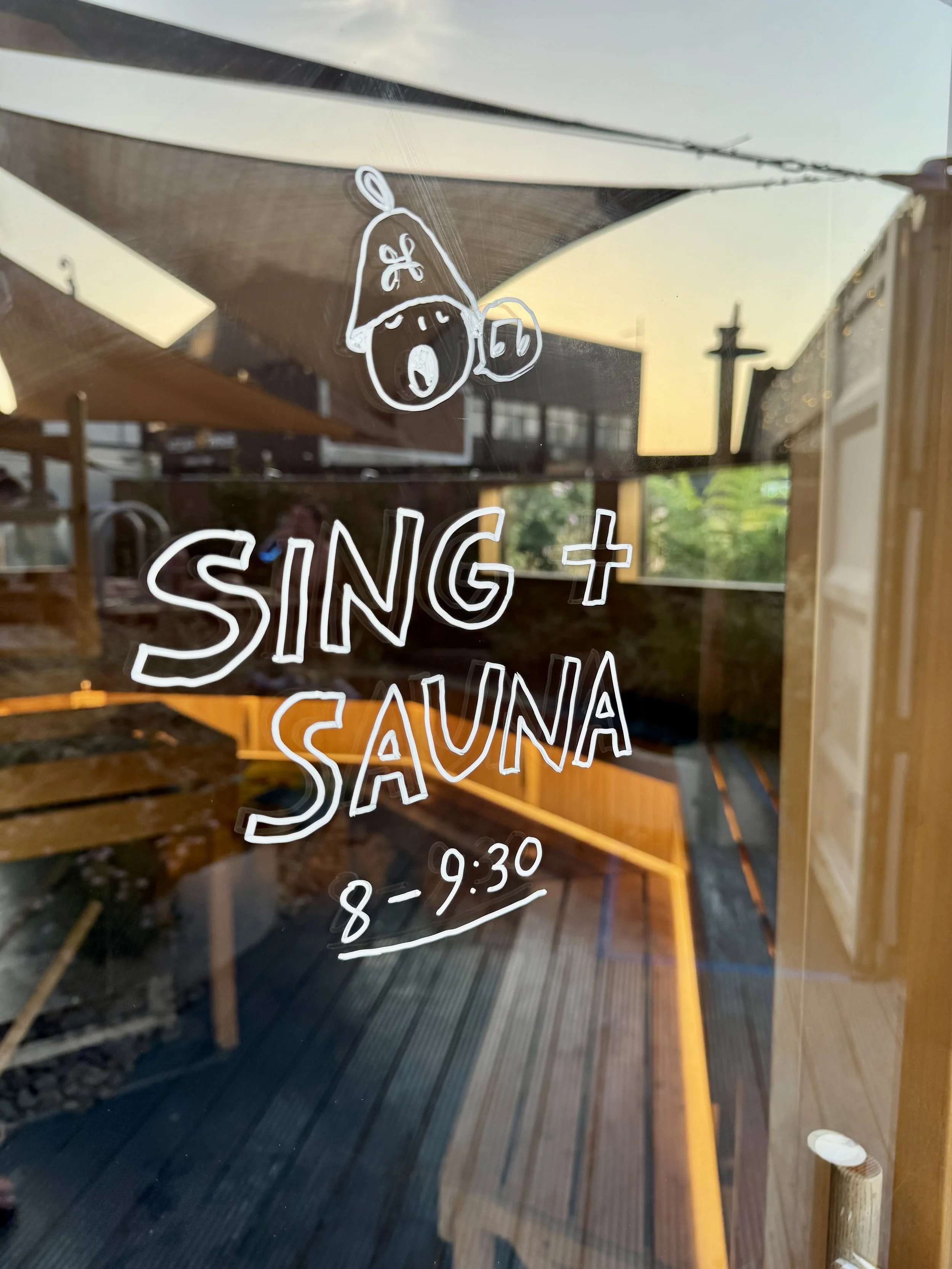 Hand-drawn 'Sing + Sauna, 8–9.30' sign on a sauna's glass door, with a singing sauna-hat character