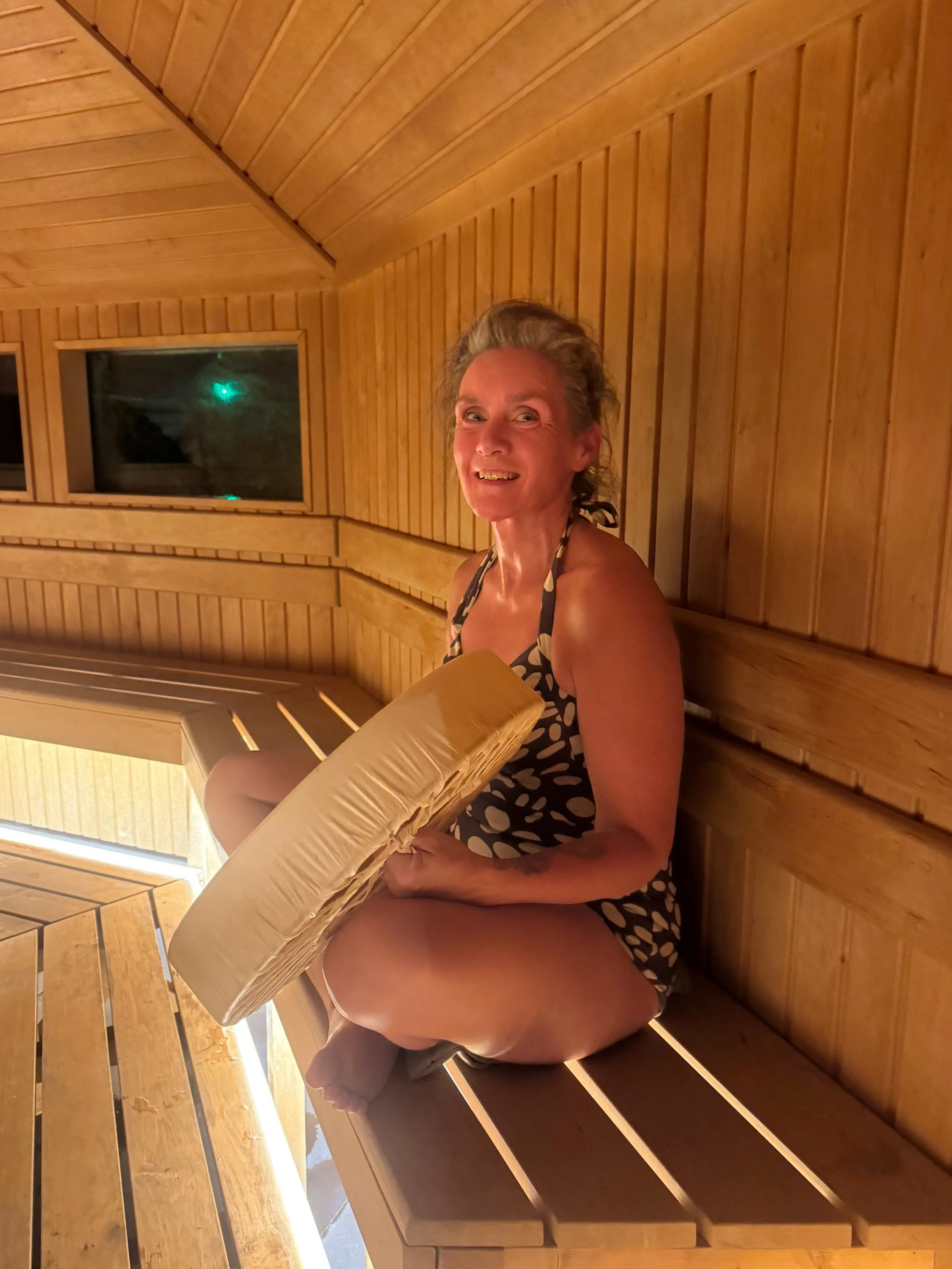 Tanja smiling in a spotted swimsuit, sitting cross-legged on a sauna bench and holding a large frame drum