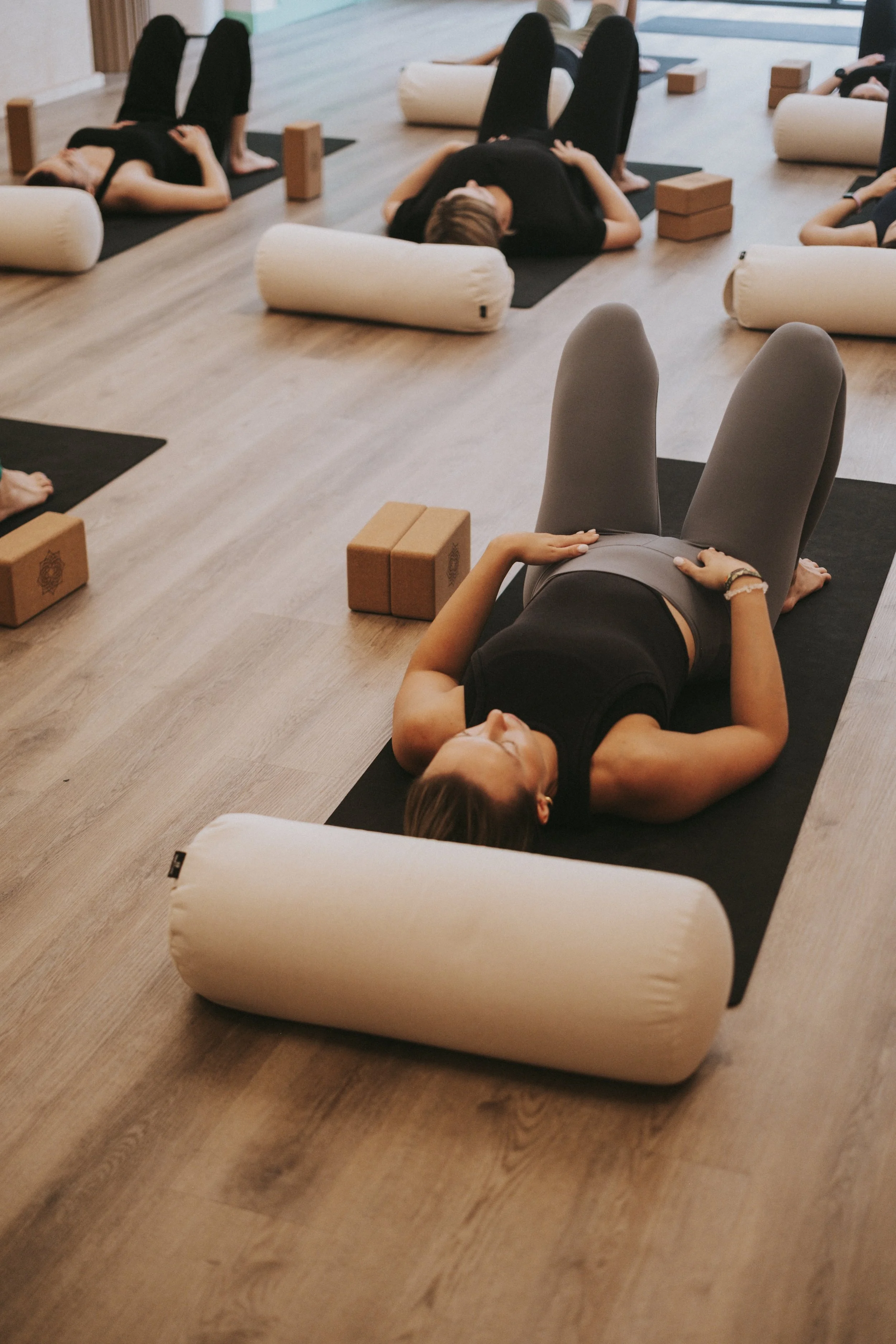 People participating in a yoga or meditation class, lying on their backs with legs bent, some with their eyes closed indoors with wooden floors, yoga props, and large windows.