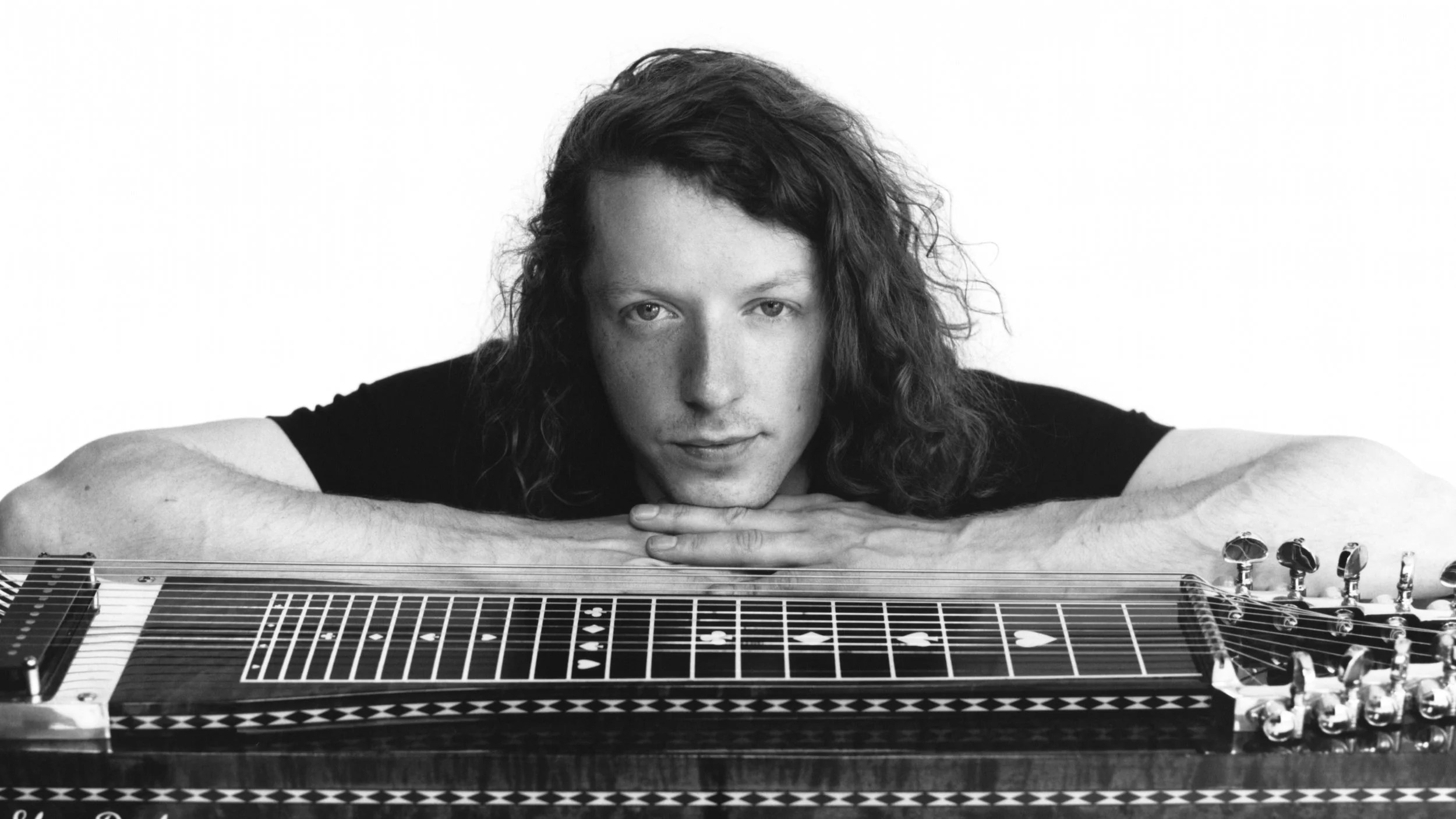 Black and white photo of a musician with long curly hair resting his chin on his arms over a pedal steel guitar