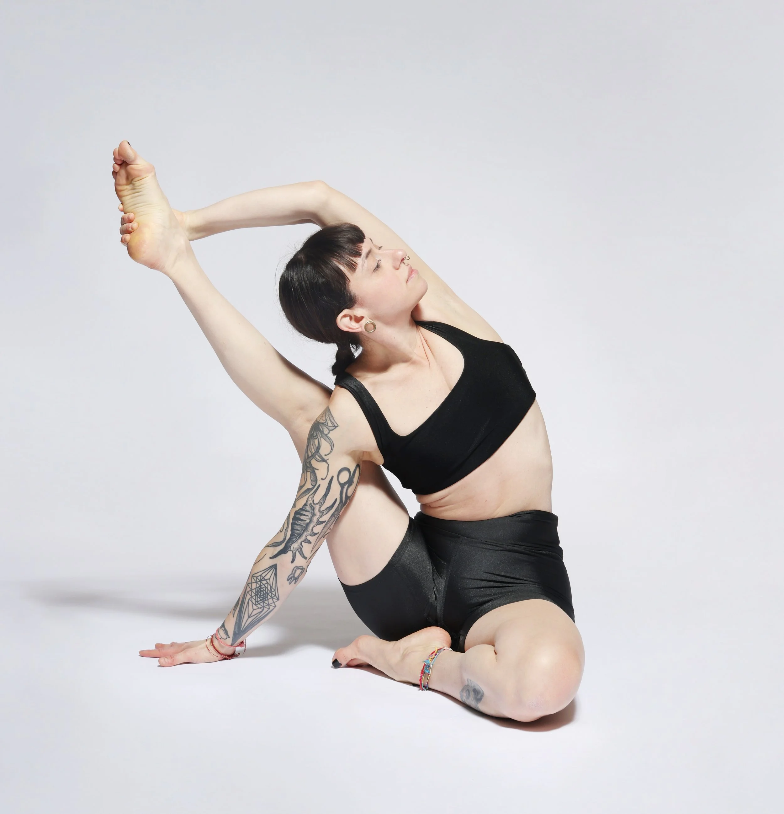 Yoga teacher Antonella in a seated twist, one arm reaching back to hold her raised foot, wearing black against a pale grey background.