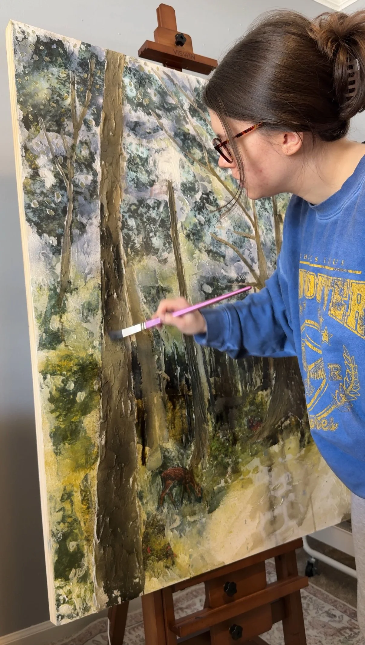 A woman with glasses painting a landscape scene of trees and a deer on a large canvas.