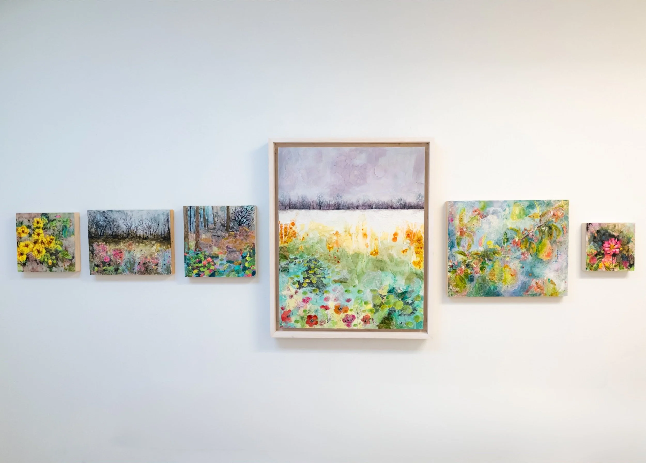 Collection of seven colorful landscape and floral paintings hung on a white gallery wall.