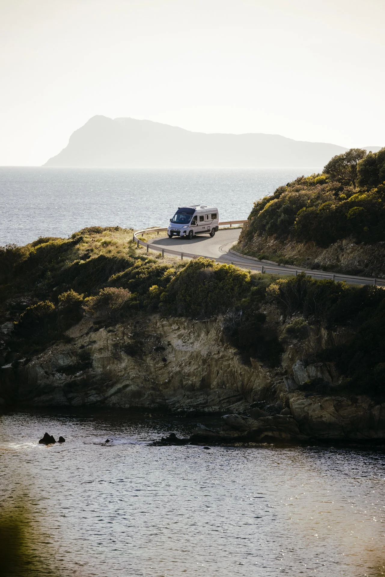 Ein Wohnmobil fährt auf einer kurvigen Küstenstraße entlang des Meeres, mit grün bewachsenen Hügeln im Vordergrund und einer Insel im Hintergrund, bei Sonnenlicht.
