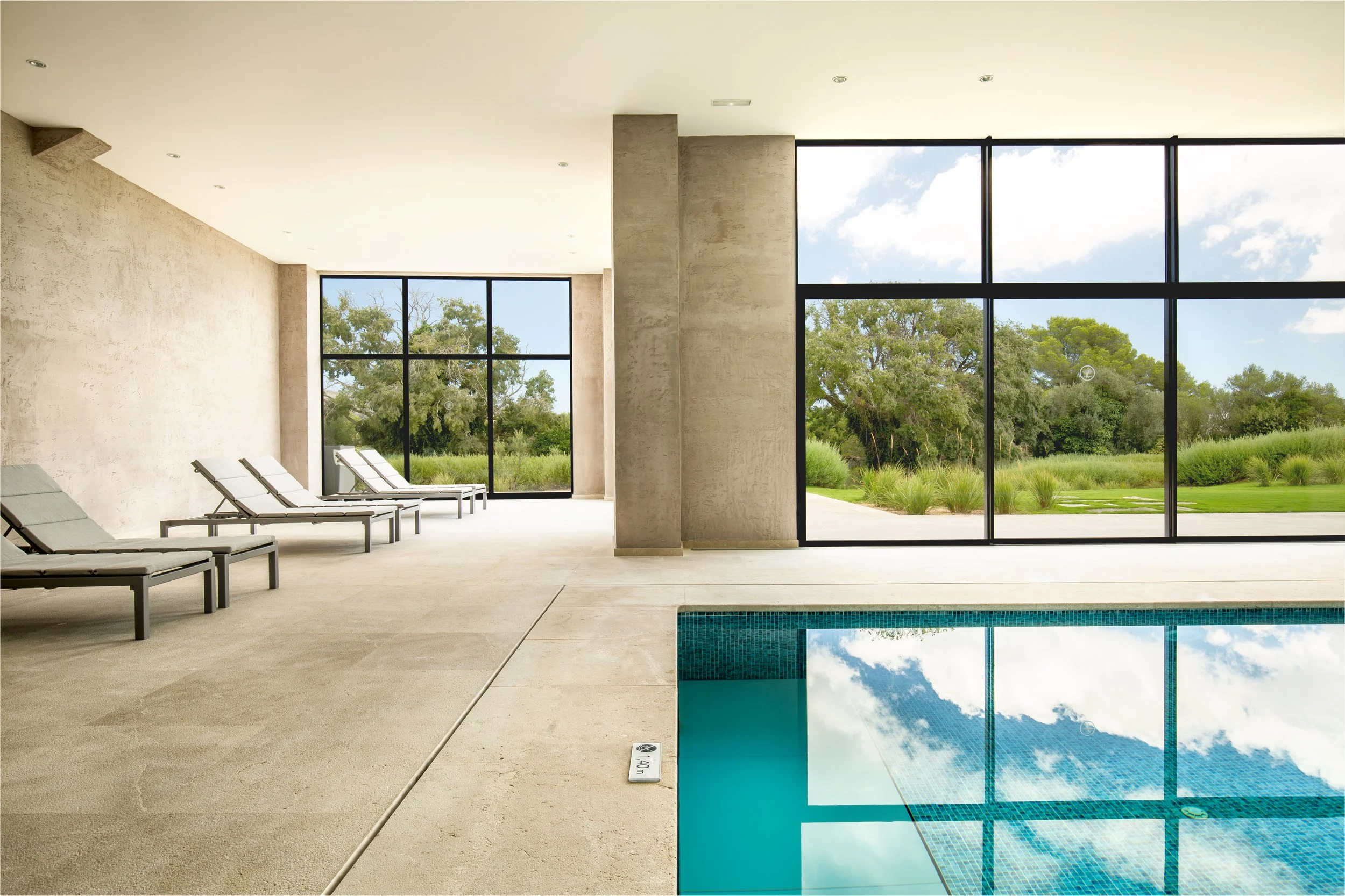 Innenaufnahme eines Spa- oder Hotelpools mit Liegen und großen Fenstern, die einen Blick auf einen Garten mit Bäumen und grünem Rasen bieten.