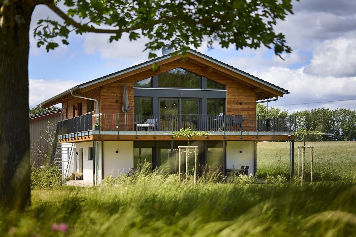 Ein modernes Haus mit Holzfassade und Balkon, umgeben von grünem Gras und Bäumen unter einem Himmel mit Wolken.