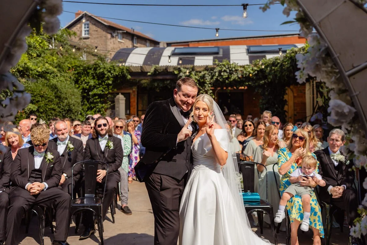 Wedding couple at the end of their ceremony outdoors at the Shack Revolution, playfully posing doing peace signs into camera, surrounded by wedding guests.