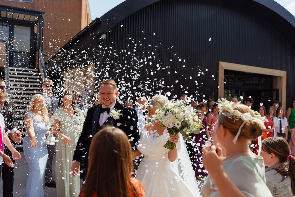Wedding couple having confetti thrown over them by wedding guests, white confetti fluttering, outside the Shack Revolution in Hereford.