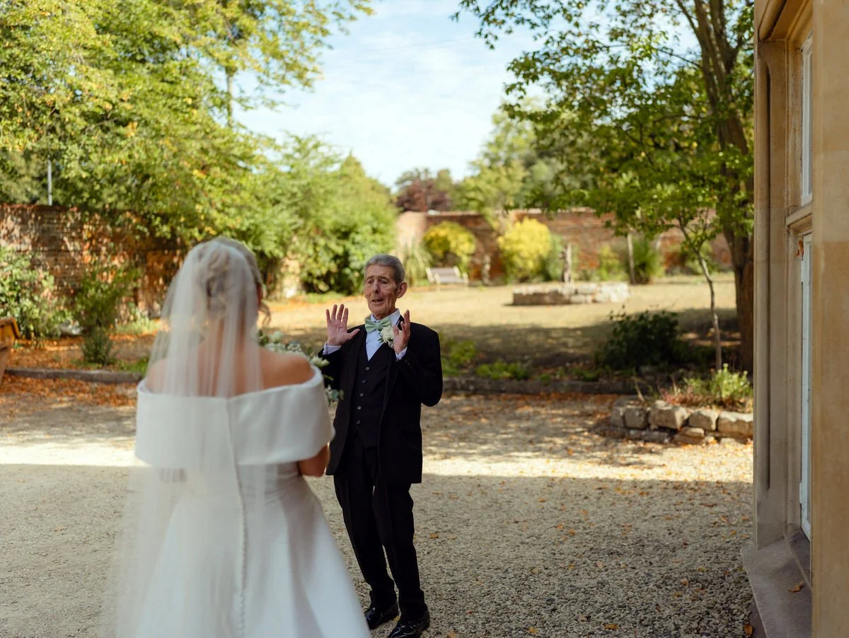 Father of the bride playing with mock shock holding his hands up at first seeing his daughter, the bride in her wedding dress.