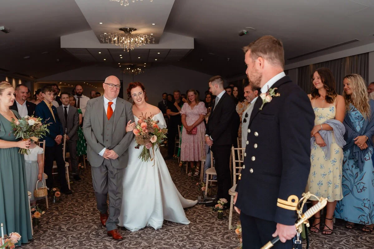 Wedding ceremony, bridge walking down the aisle with her father, clutching her bouquet inside the Bear Hotel in Cowbridge.