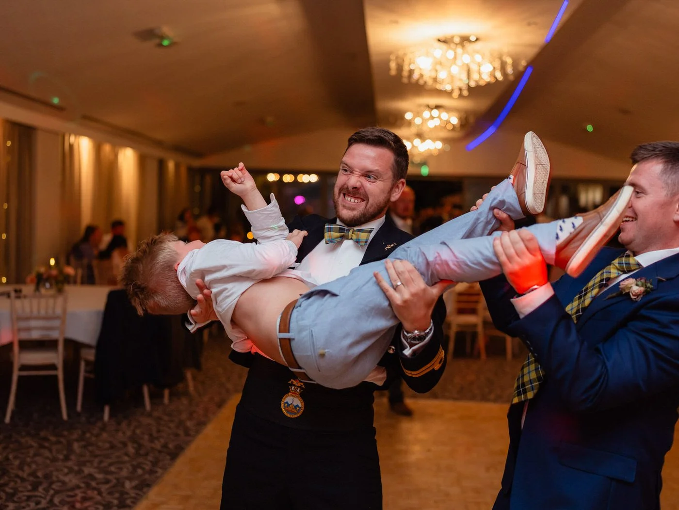 Male groom on his wedding day on the dancefloor having fun holding up a small child in the air.