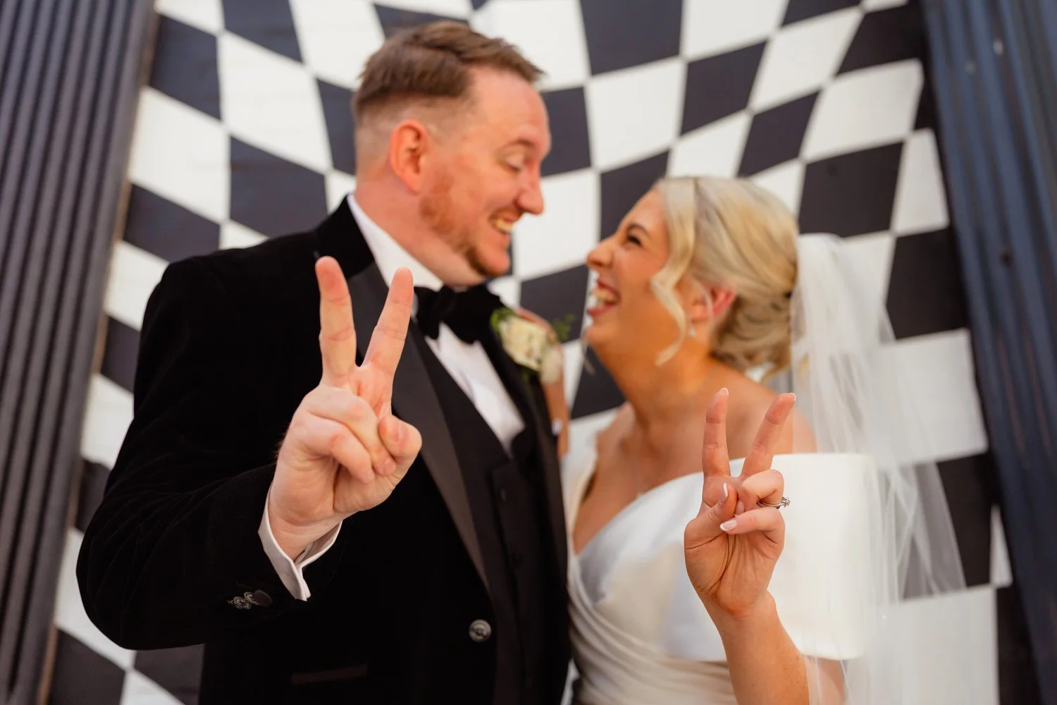 Wedding couple against a black and white checked wall outside Shack Revolution in Hereford. Male and female wedding couple doing the peace sign and looking at each other laughing.