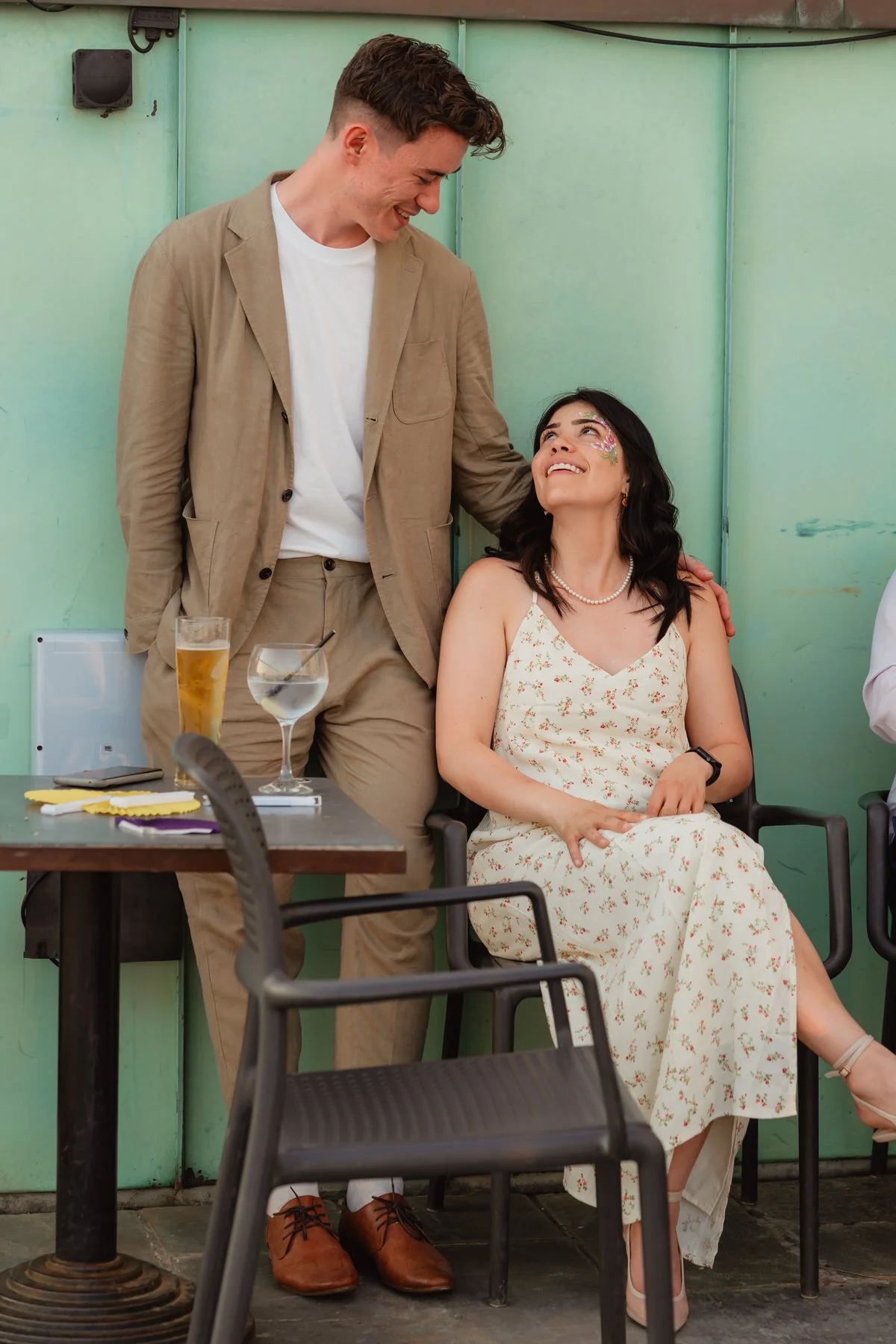 Two wedding guests looking at each other with affection, a turqoise background behind them. Sat outside in the sun at the Swansea Observatory tower.