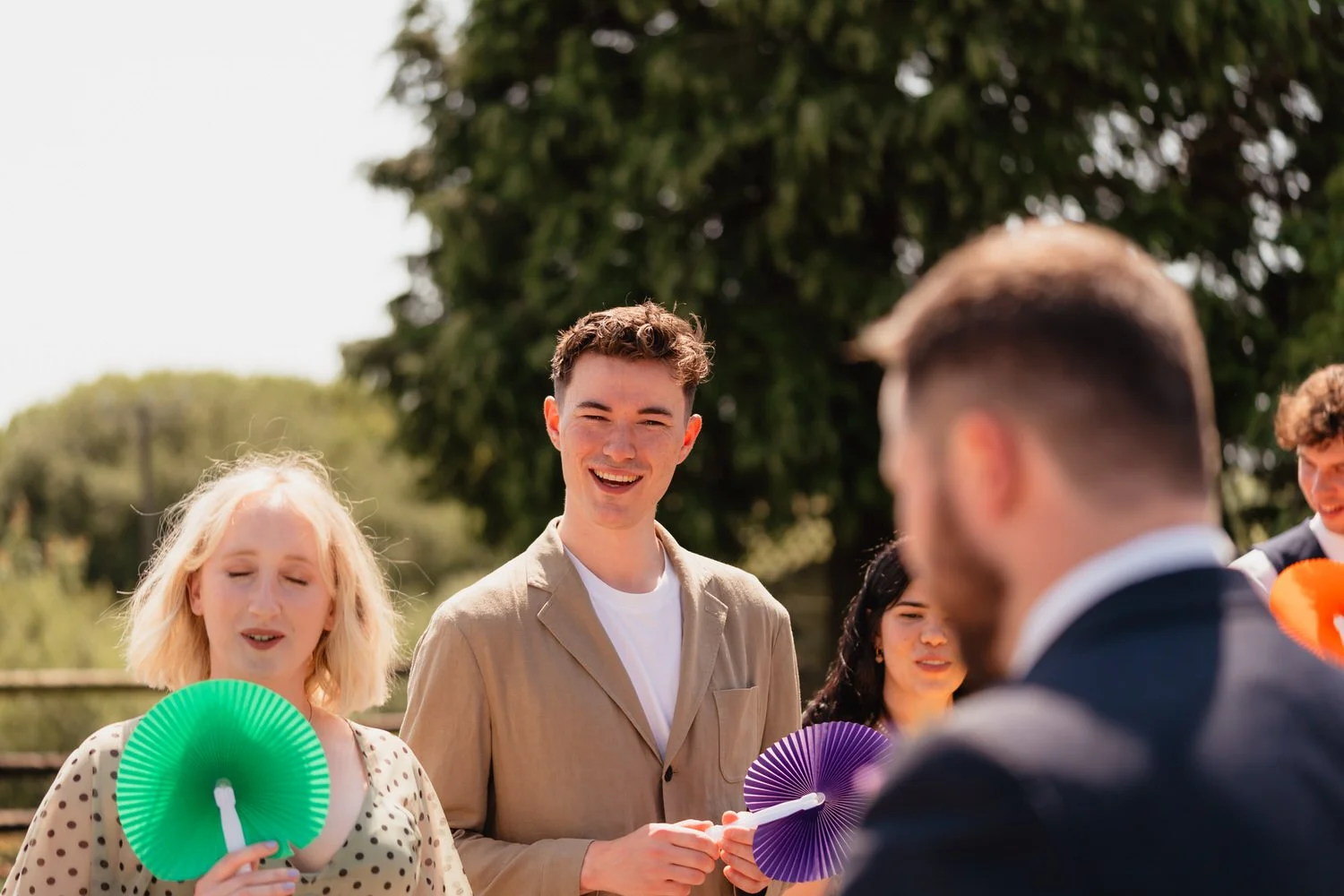 Wedding guests enjoying themselves in the sun outside Clyne Farm, holding coloured fans.
