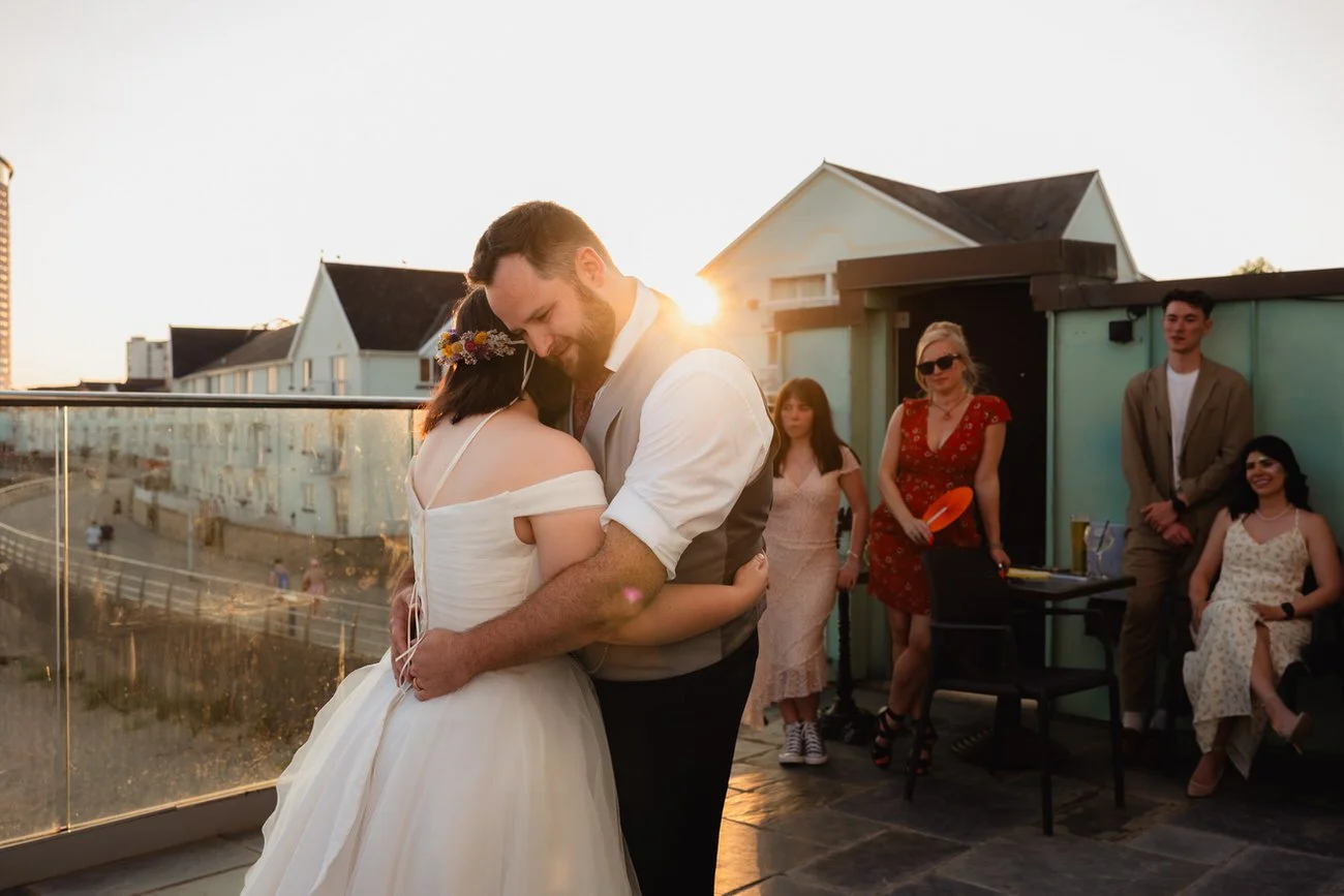 Wedding couple celebrating their vows dancing during their first dance with the sun setting behind them, sun flare showing, guests watching on behind them, balcony showing view on to Swansea beach, situated at Swansea Observatory Tower.