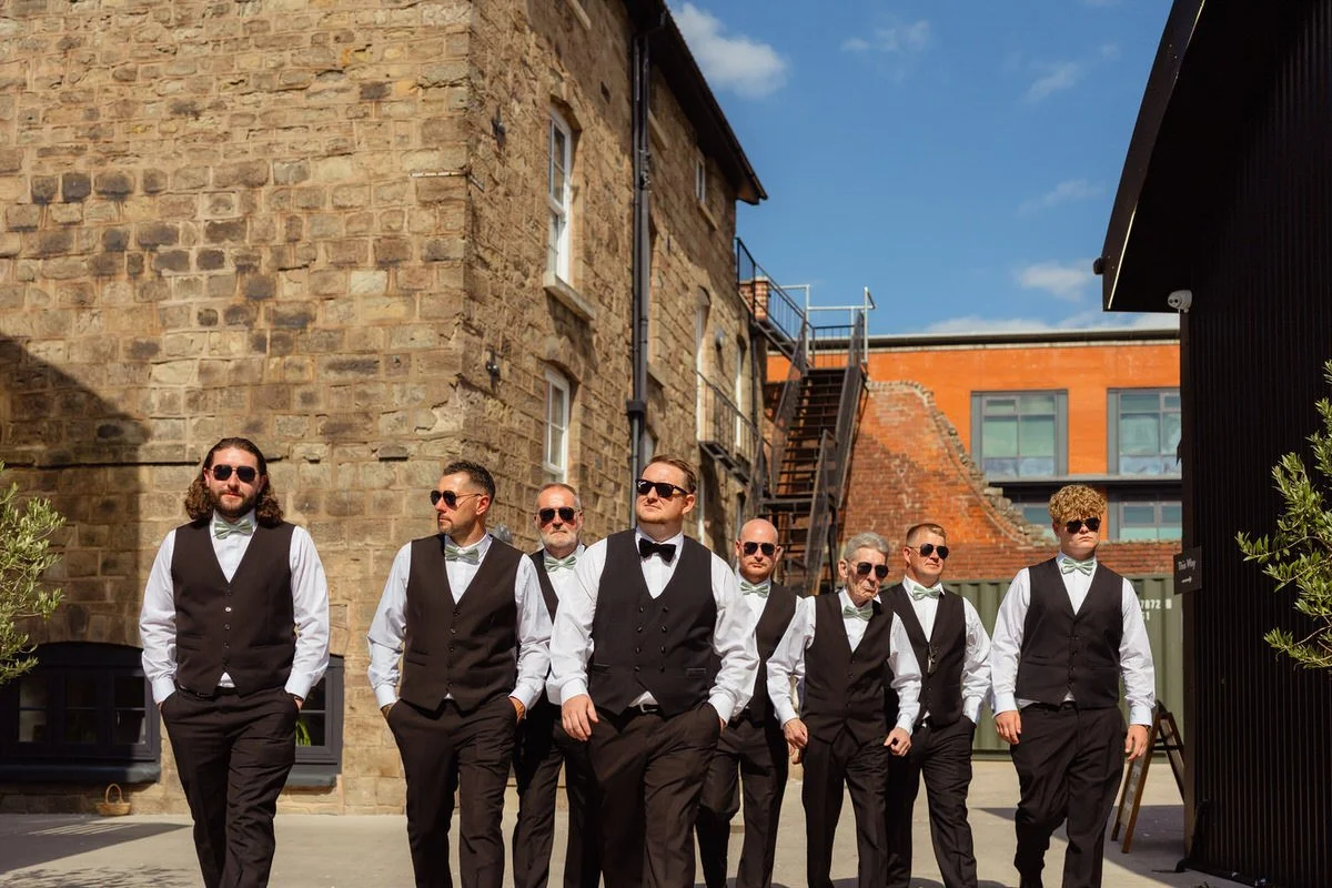 Group of the male wedding party in matching suits, bow ties, and dark sunglasses on, all walking outside Shack Revolution.