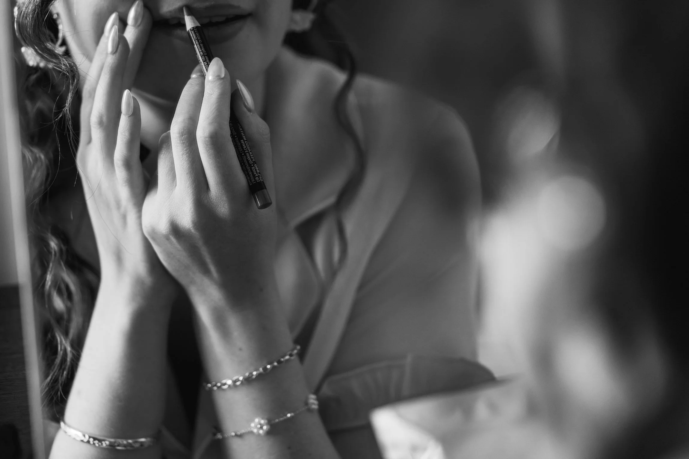 A woman applies lip liner in front of a mirror, her hands and face partially visible in black and white, bracelets on her wrists. .