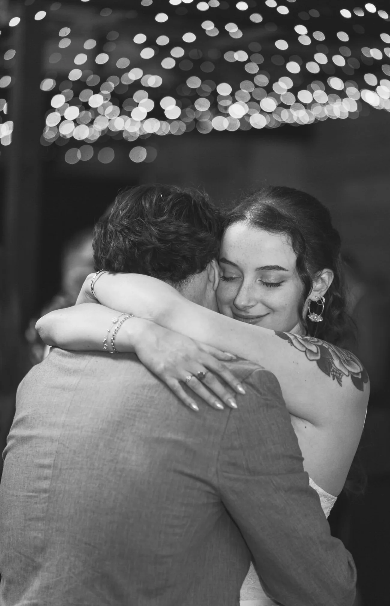 Black and white first dance photo, womans face seen smiling calmly, her arms wrapped around her dance partner, out of focus bokeh lights in the background at the Shack Revolution wedding in Hereford.