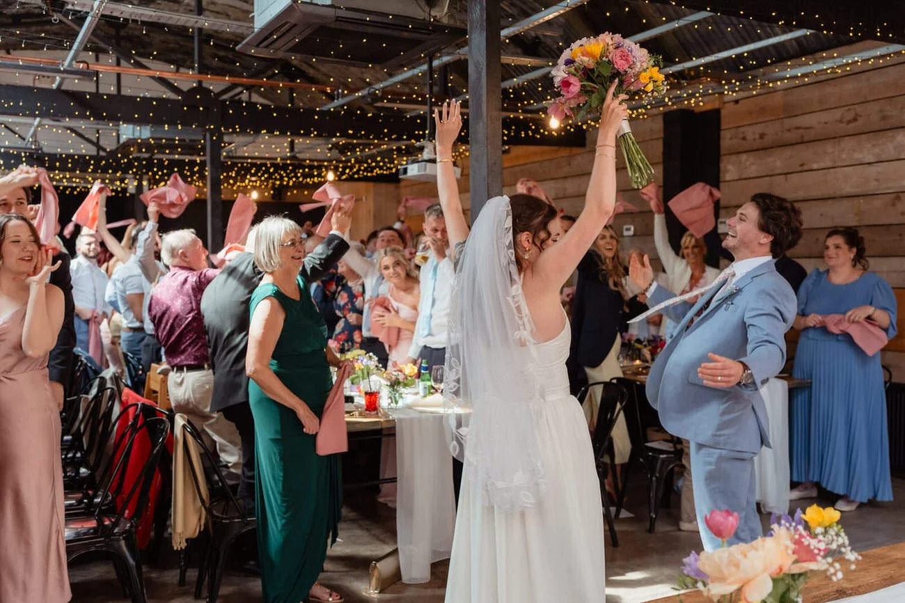 Lively wedding breakfast entrance, guests all on their feet waving napkins, wedding couple excited celebrating with their hands in the air, inside the Shack Revolution in Hereford.