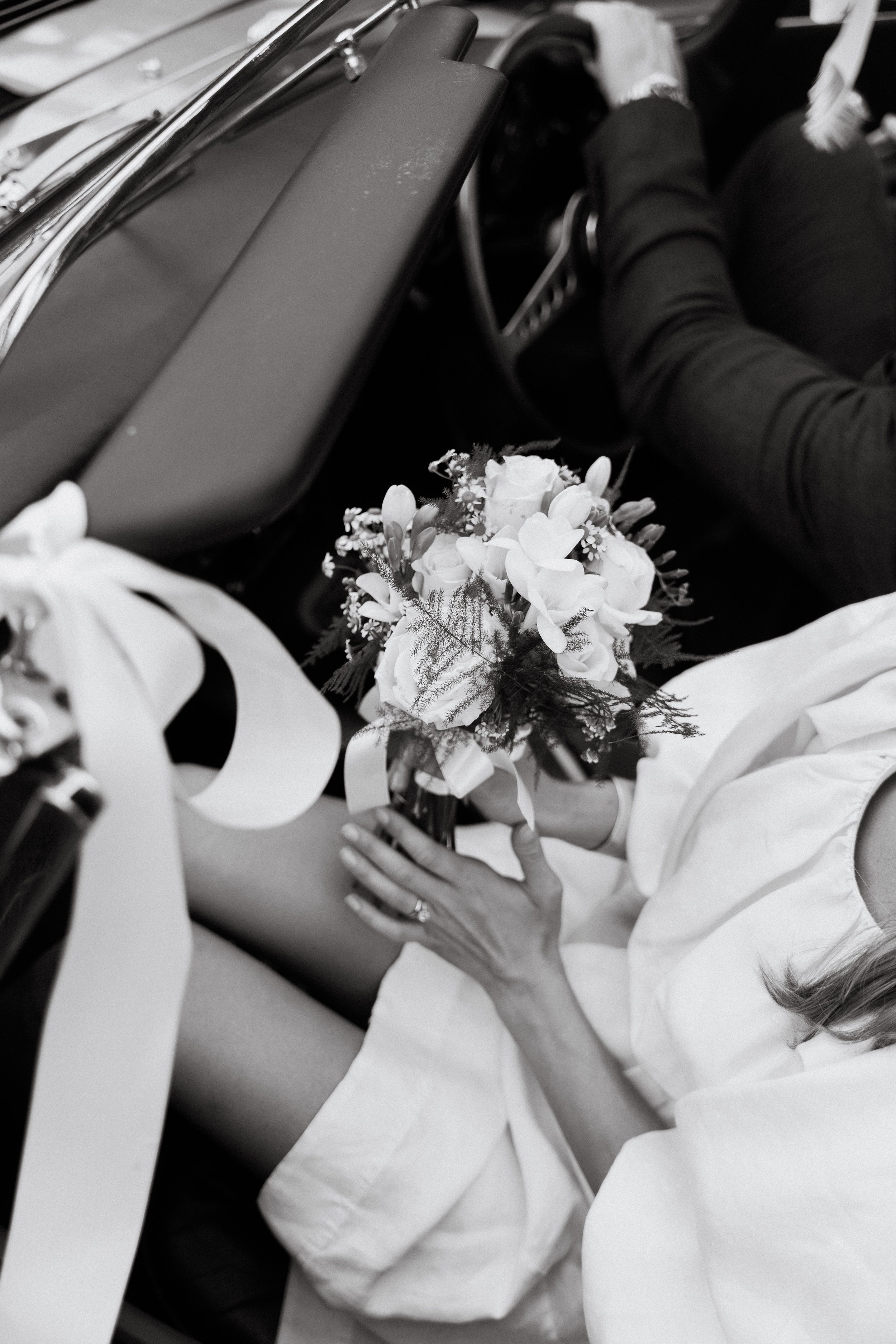A bride holding a bouquet of flowers inside a car, seated with her legs exposed and wearing a white dress. Wedding decorations, including ribbons, are visible.