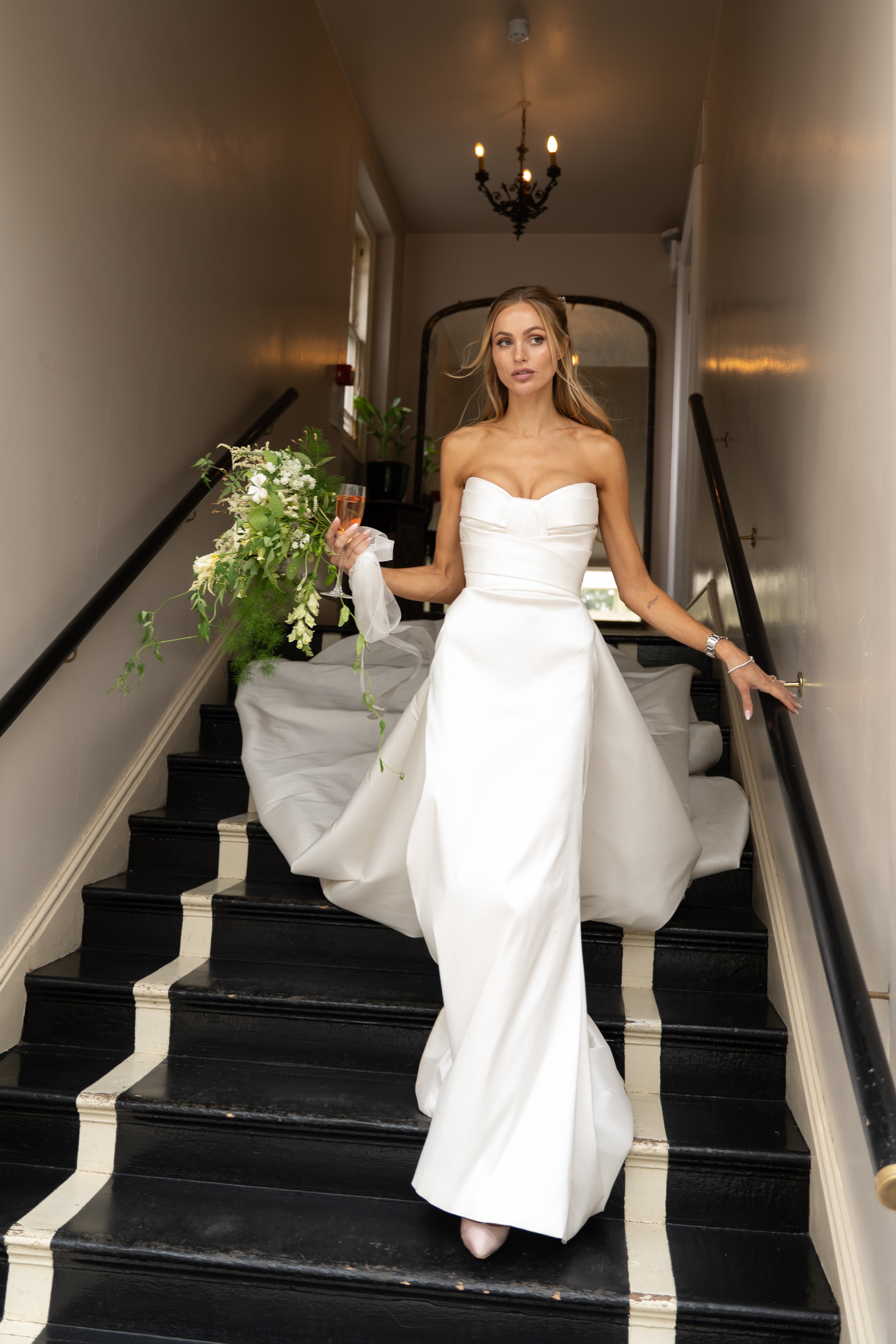 A bride in a strapless white wedding dress holding a bouquet and a glass of champagne, walking down the stairs of a building.