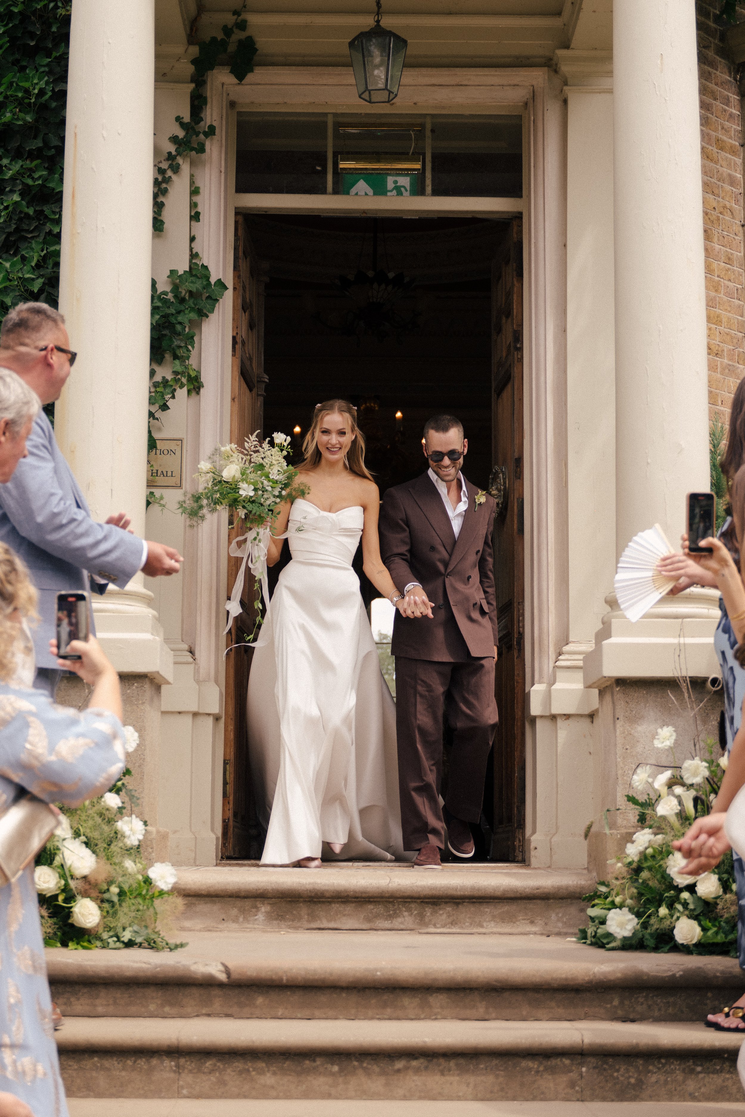 A newly married couple, the bride in a white strapless wedding gown holding a bouquet, and the groom in a brown suit, smiling as they walk out of a church surrounded by wedding guests.