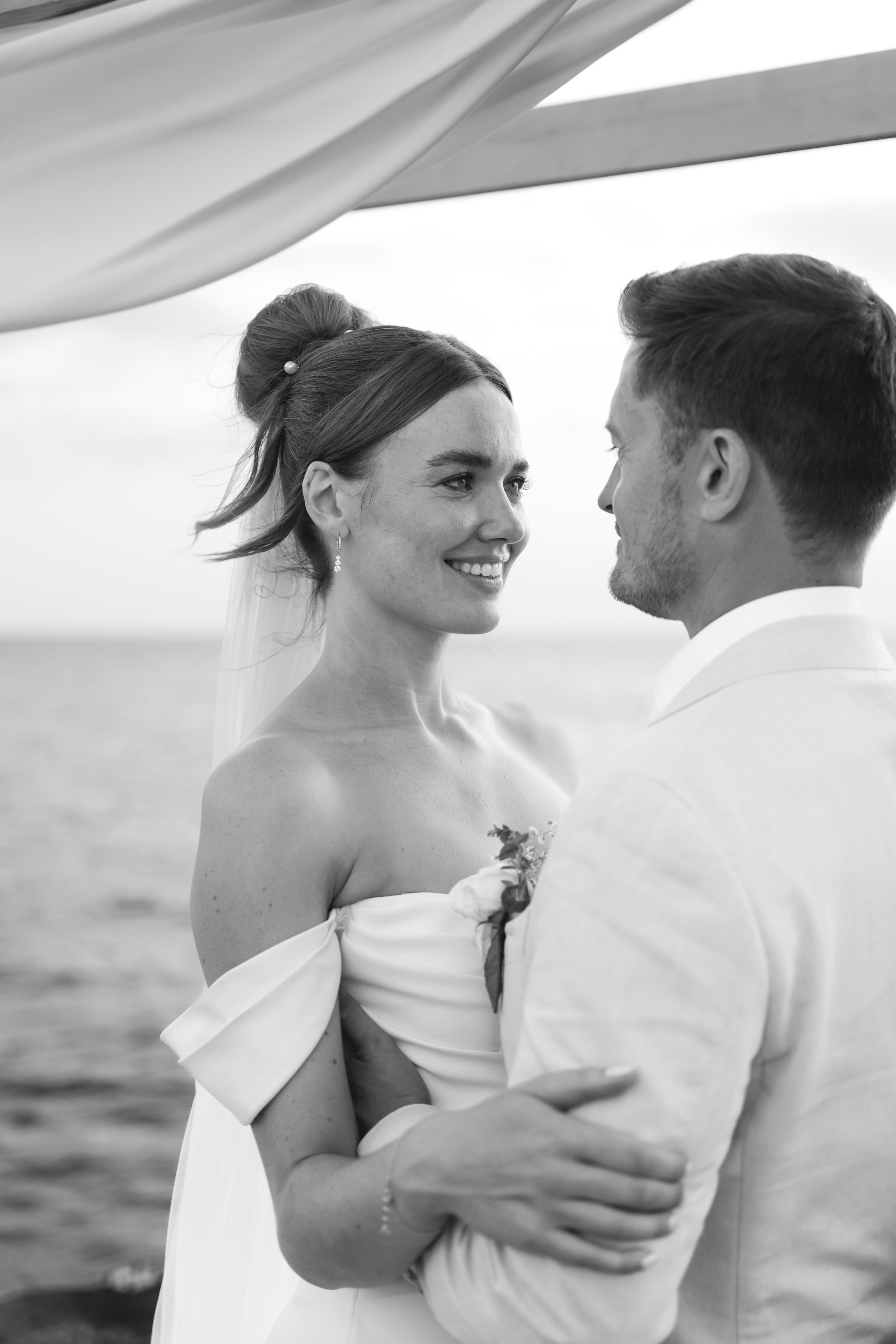 A black and white photo of a bride and groom standing close, facing each other, holding hands, with a body of water in the background during their wedding ceremony.