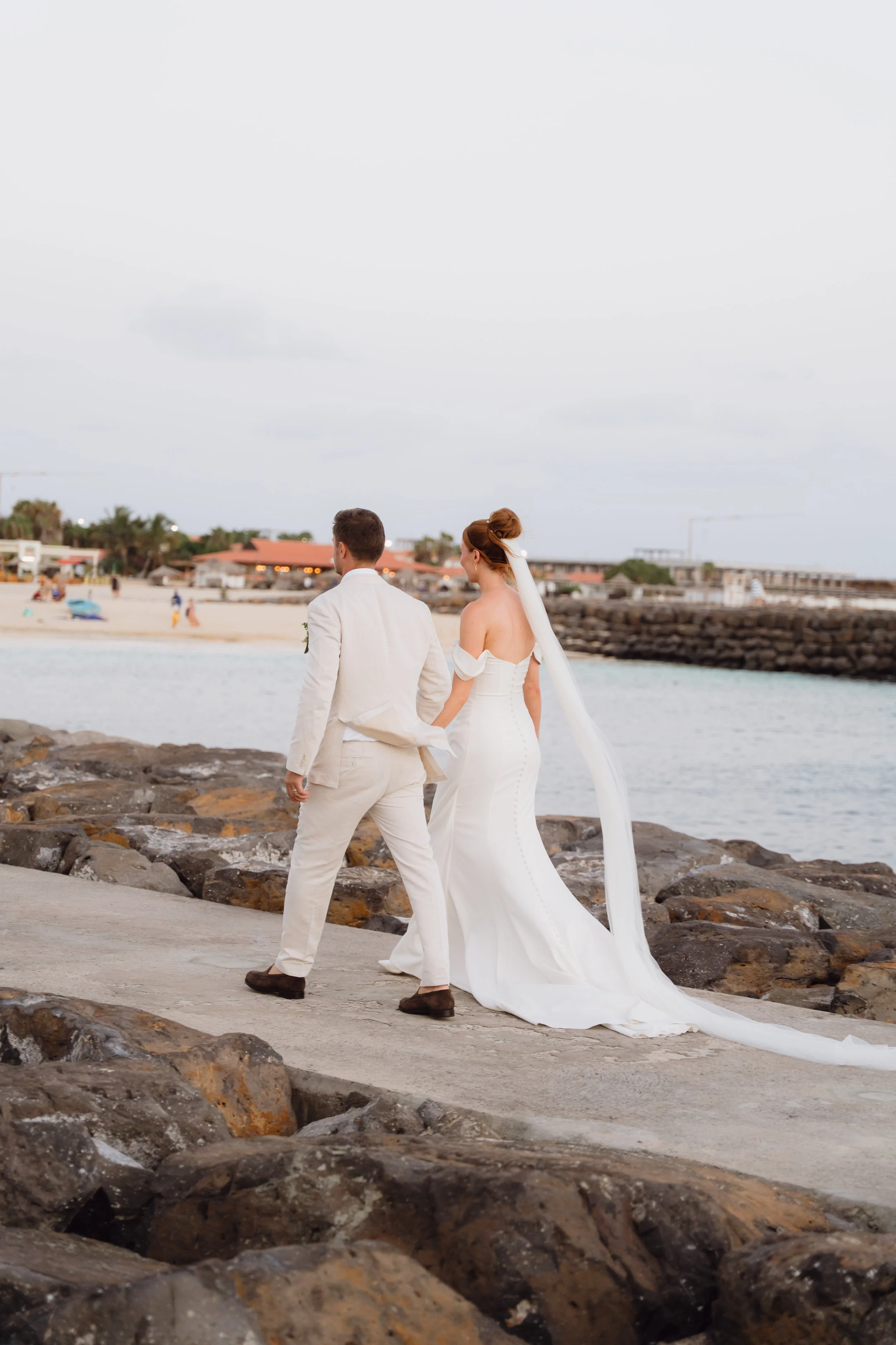 A bride and groom holding hands walking on a rocky beach in wedding attire near the water.