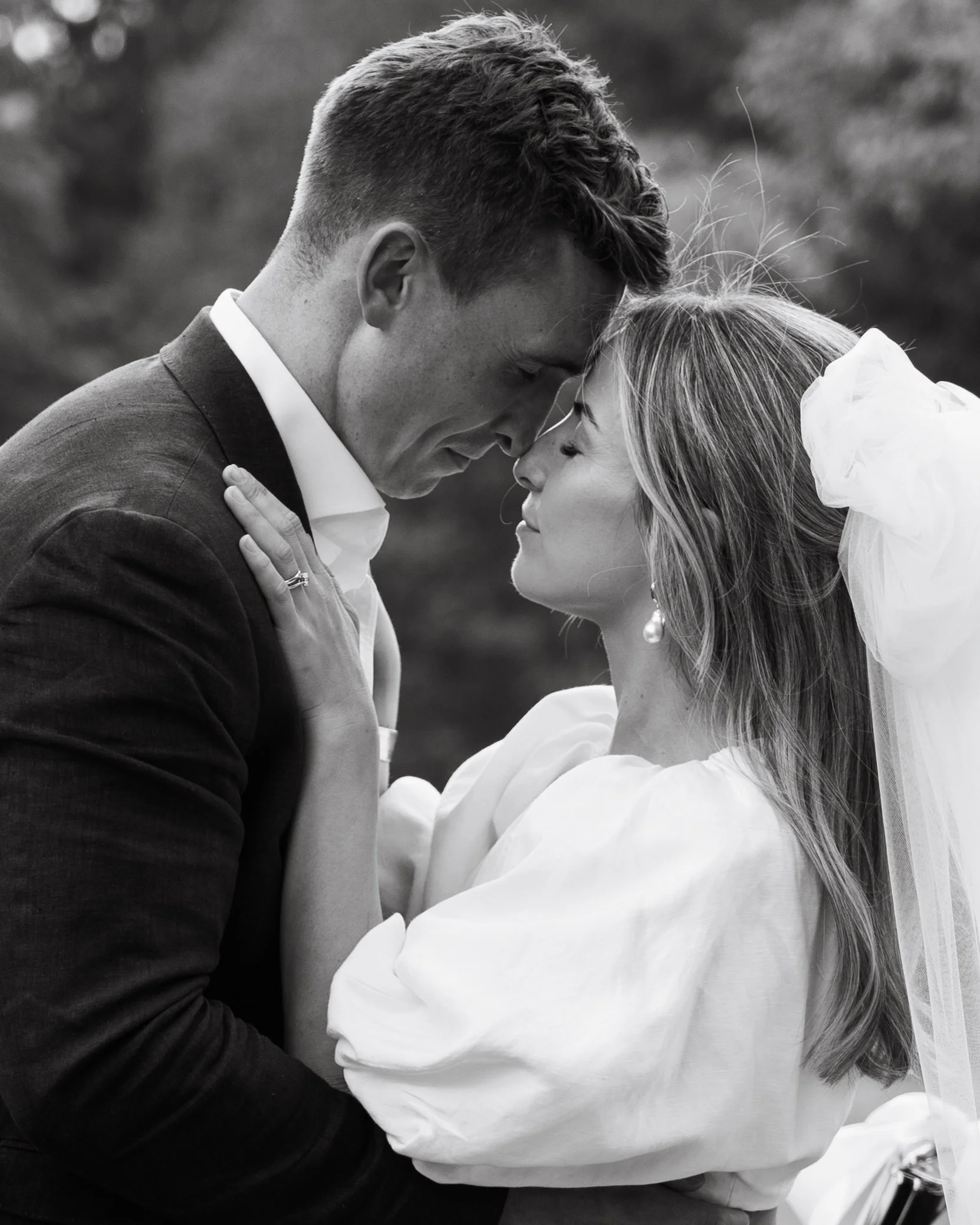 A black and white photo of a couple on their wedding day, touching foreheads and holding each other closely outdoors.